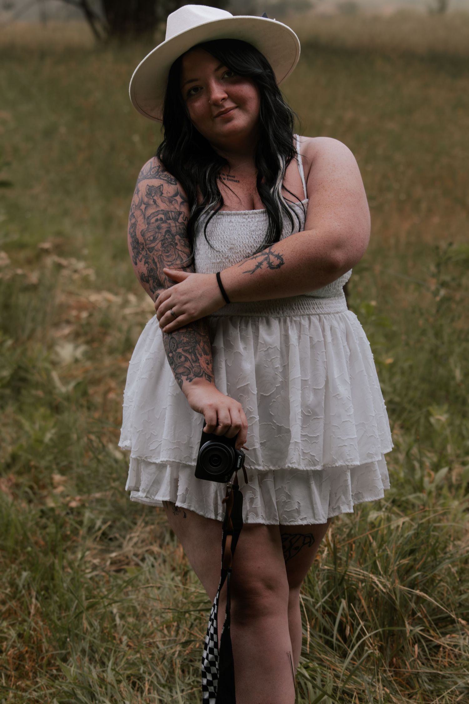 Woman in white ruffled dress and hat with tattoos holds camera in grassy field.