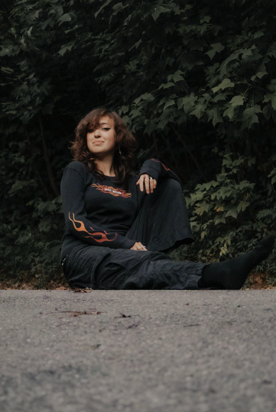 A person wearing a black sweater with flame details lounging in a natural outdoor setting beside dark foliage.