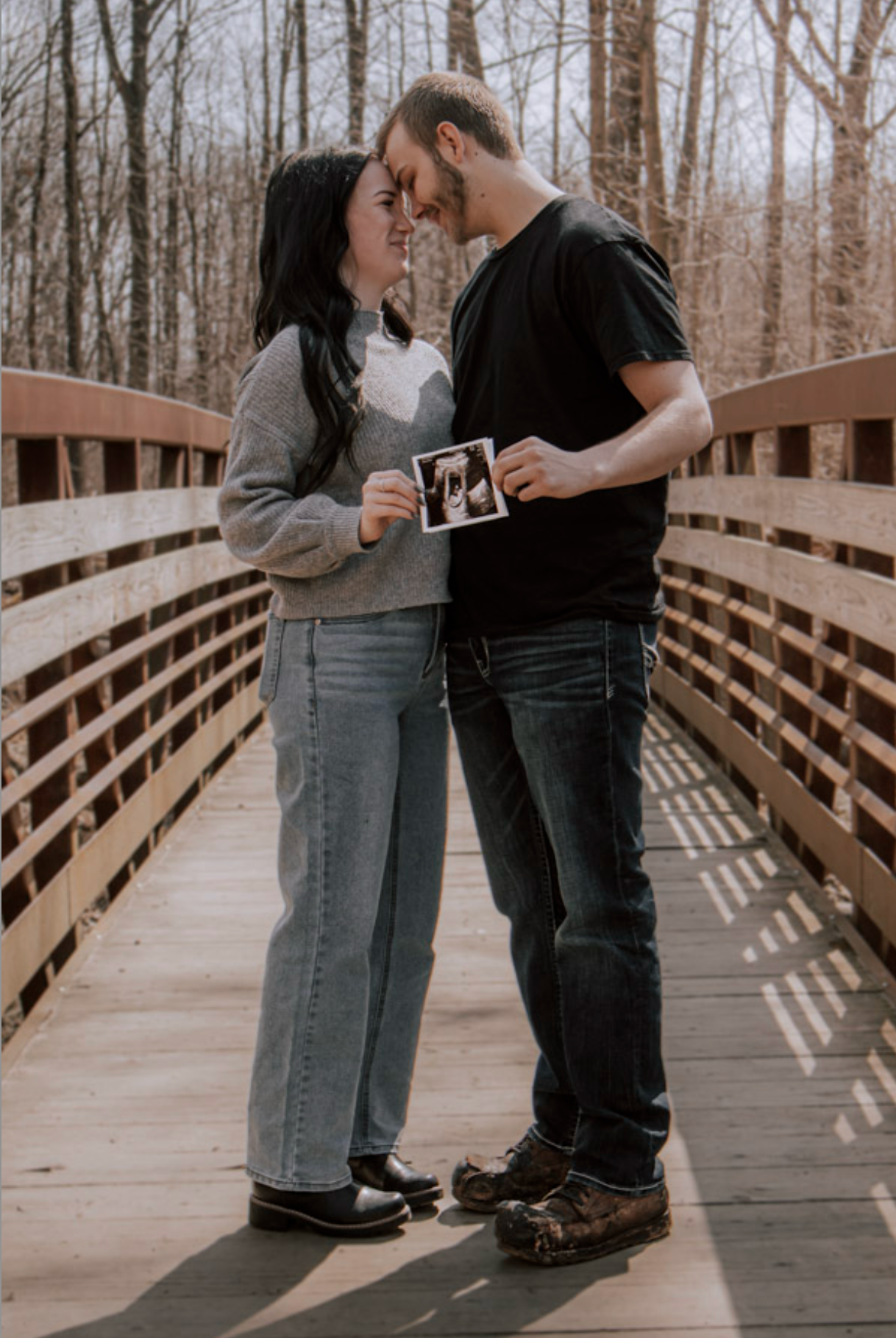 Couple sharing intimate moment on wooden bridge holding ultrasound photo announcing pregnancy.