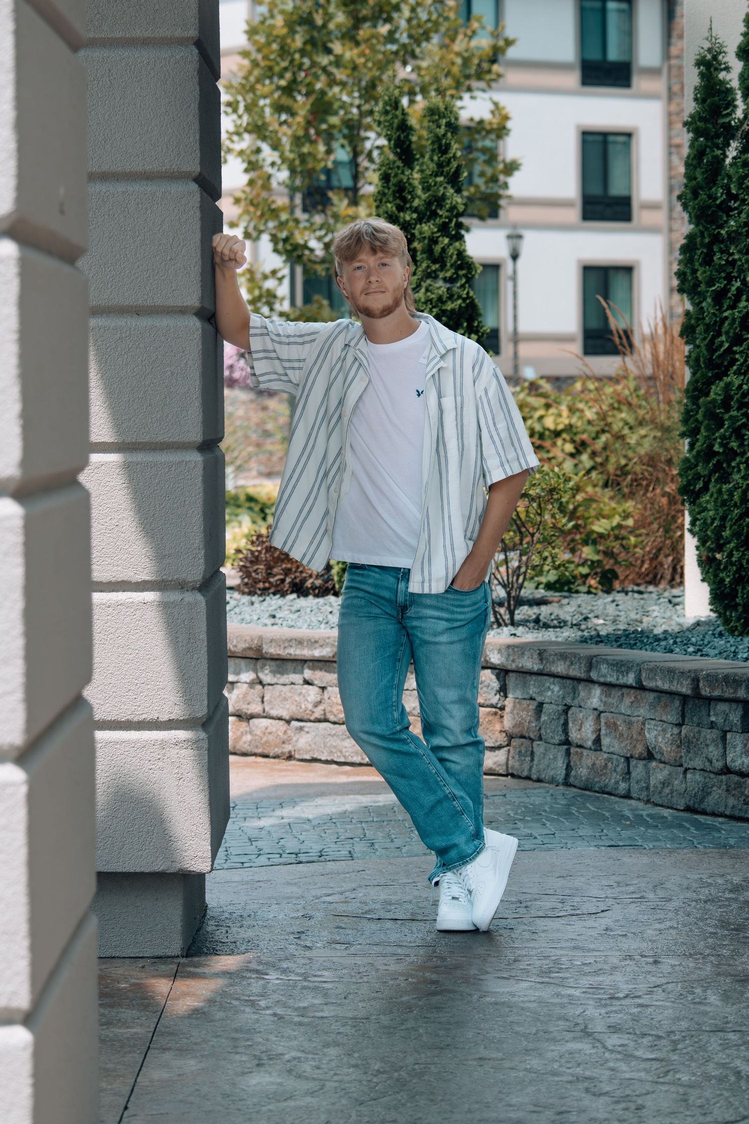 Young man in striped shirt, white tee, jeans, and sneakers posing outdoors near stone pillar with greenery.