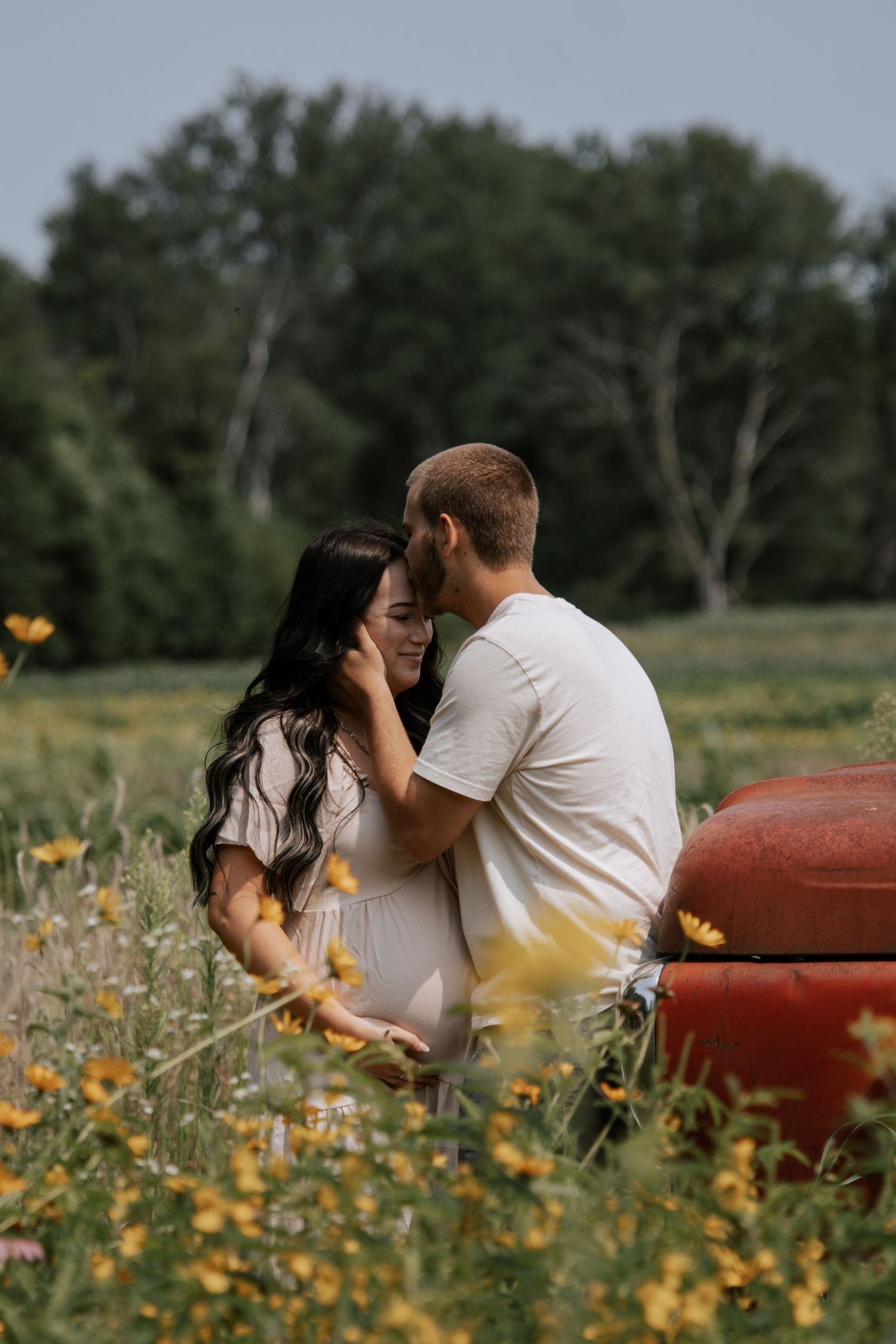 Couple sharing a romantic moment in a wildflower field near a red truck, surrounded by lush greenery.