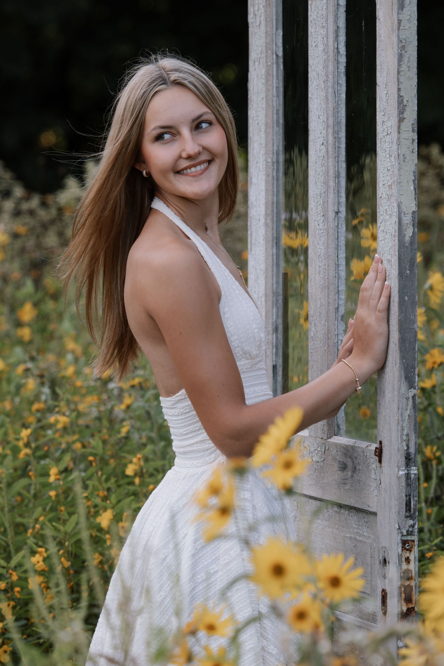 Young woman in white halter dress smiling beside rustic wooden fence surrounded by yellow wildflowers in a garden.