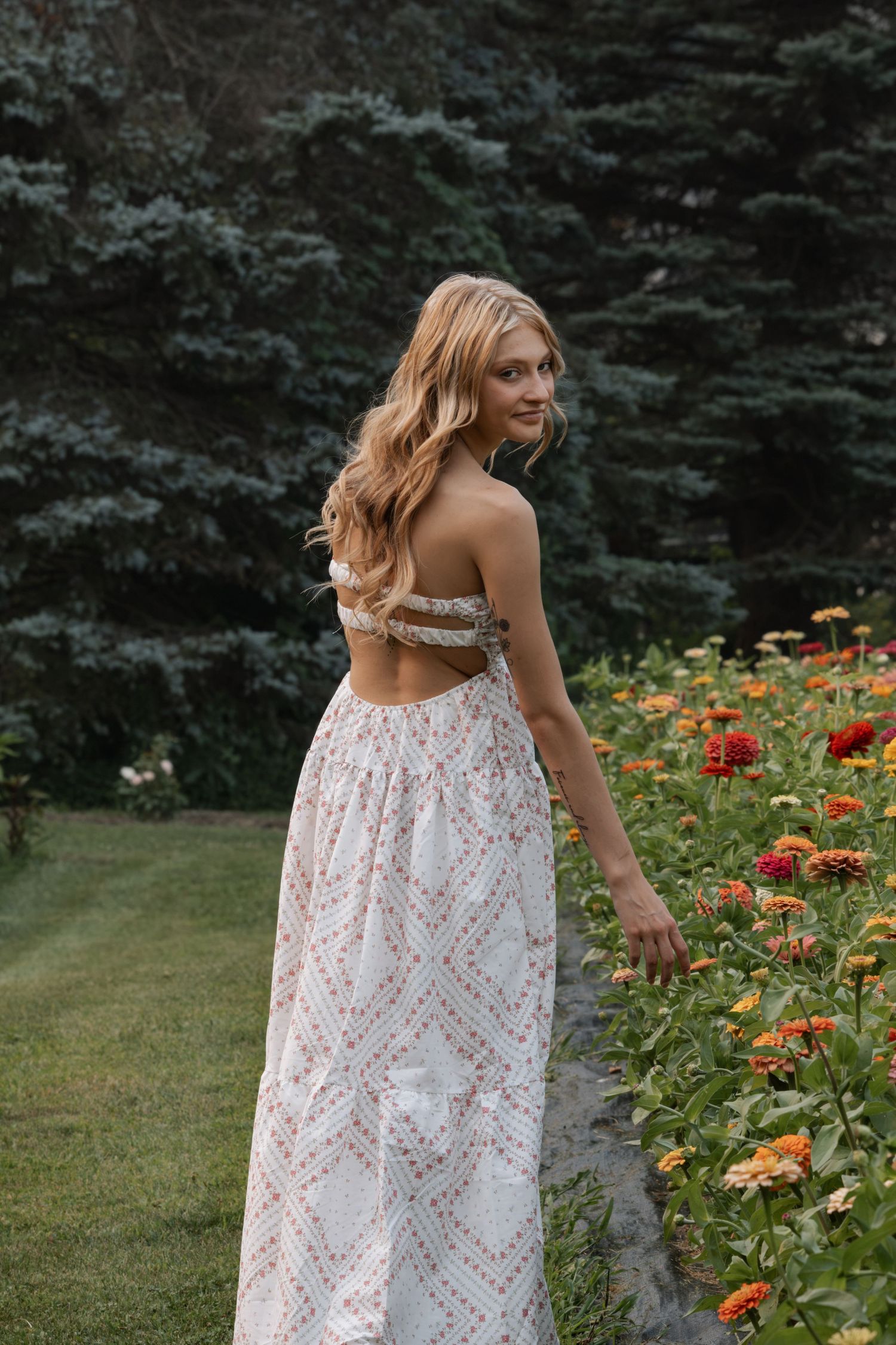 Young woman in a floral backless maxi dress standing beside colorful zinnia flowers in a lush garden.