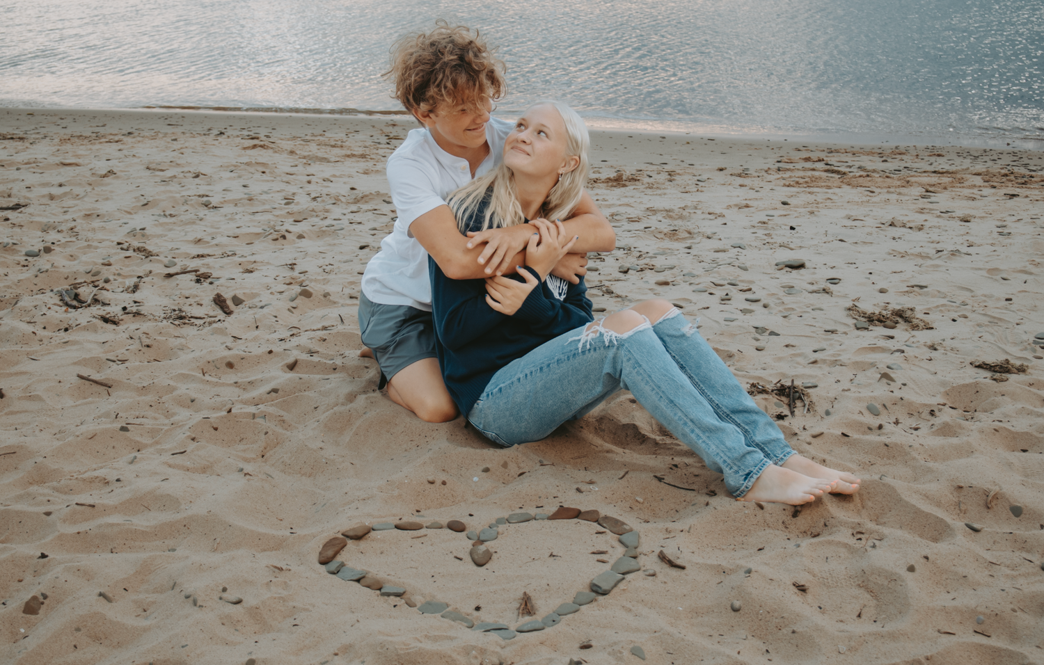 Couple sits on beach sand with heart drawn near them while enjoying romantic moment at sunset.