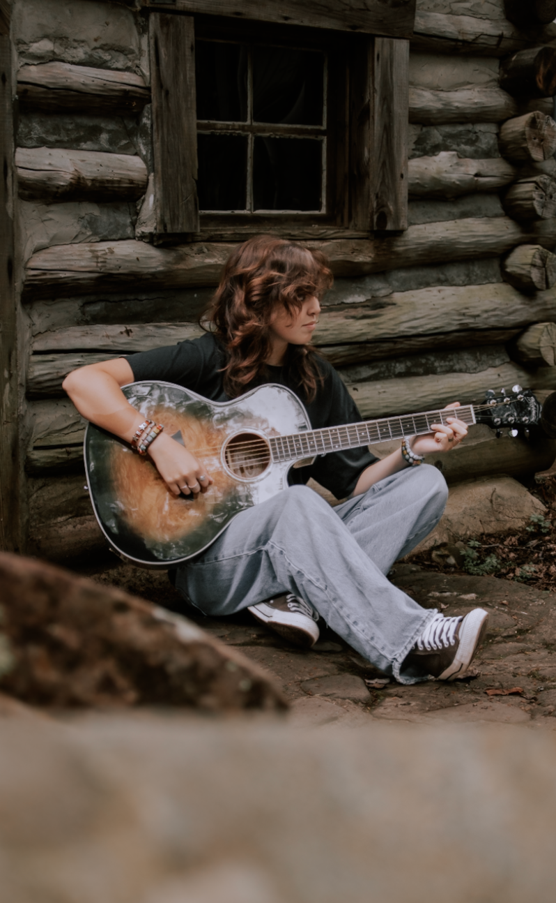 A rustic log cabin scene with a musician playing acoustic guitar while relaxing on the ground.