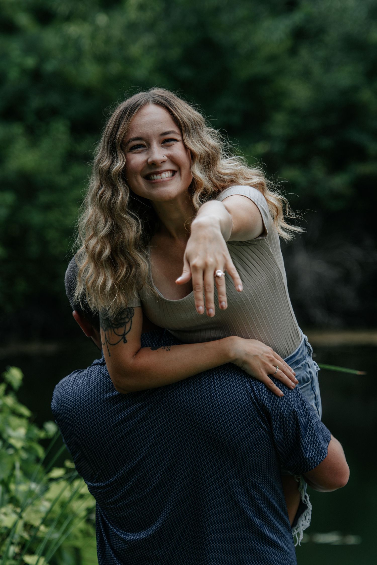 Smiling woman showing engagement ring while getting a piggyback ride outdoors near lush greenery.