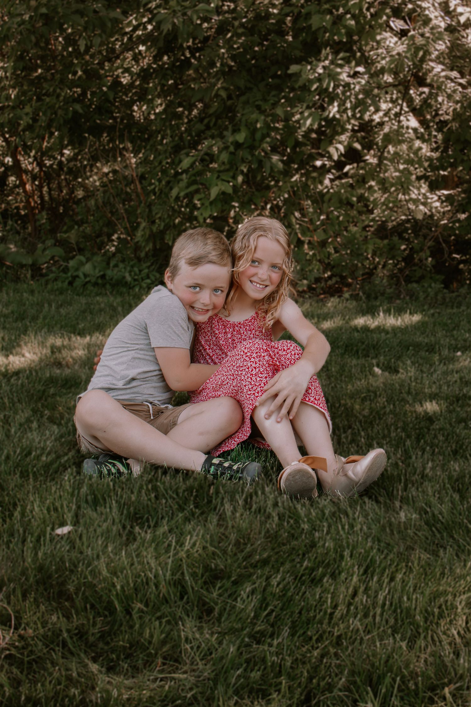 Two young siblings hugging and smiling on green grass outdoors, boy in gray shirt and girl in red floral dress.