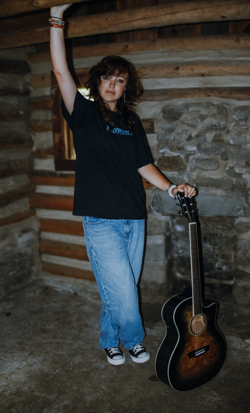 Casual musician in black t-shirt and jeans poses with acoustic guitar against rustic stone wall in moody lighting.