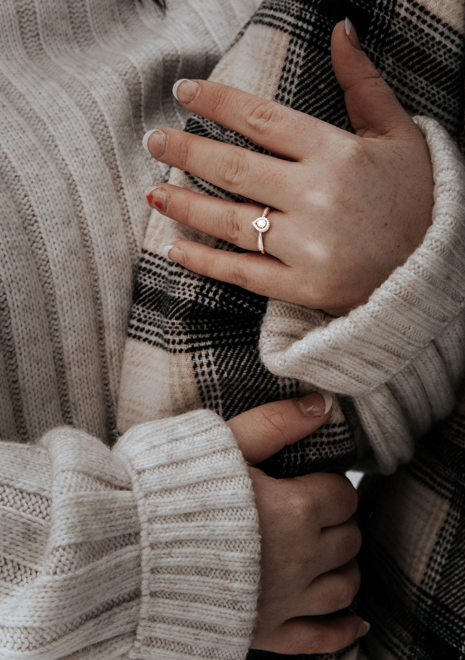 Close-up photo of hands wearing a white knit sweater and delicate engagement ring against a textured gray background.