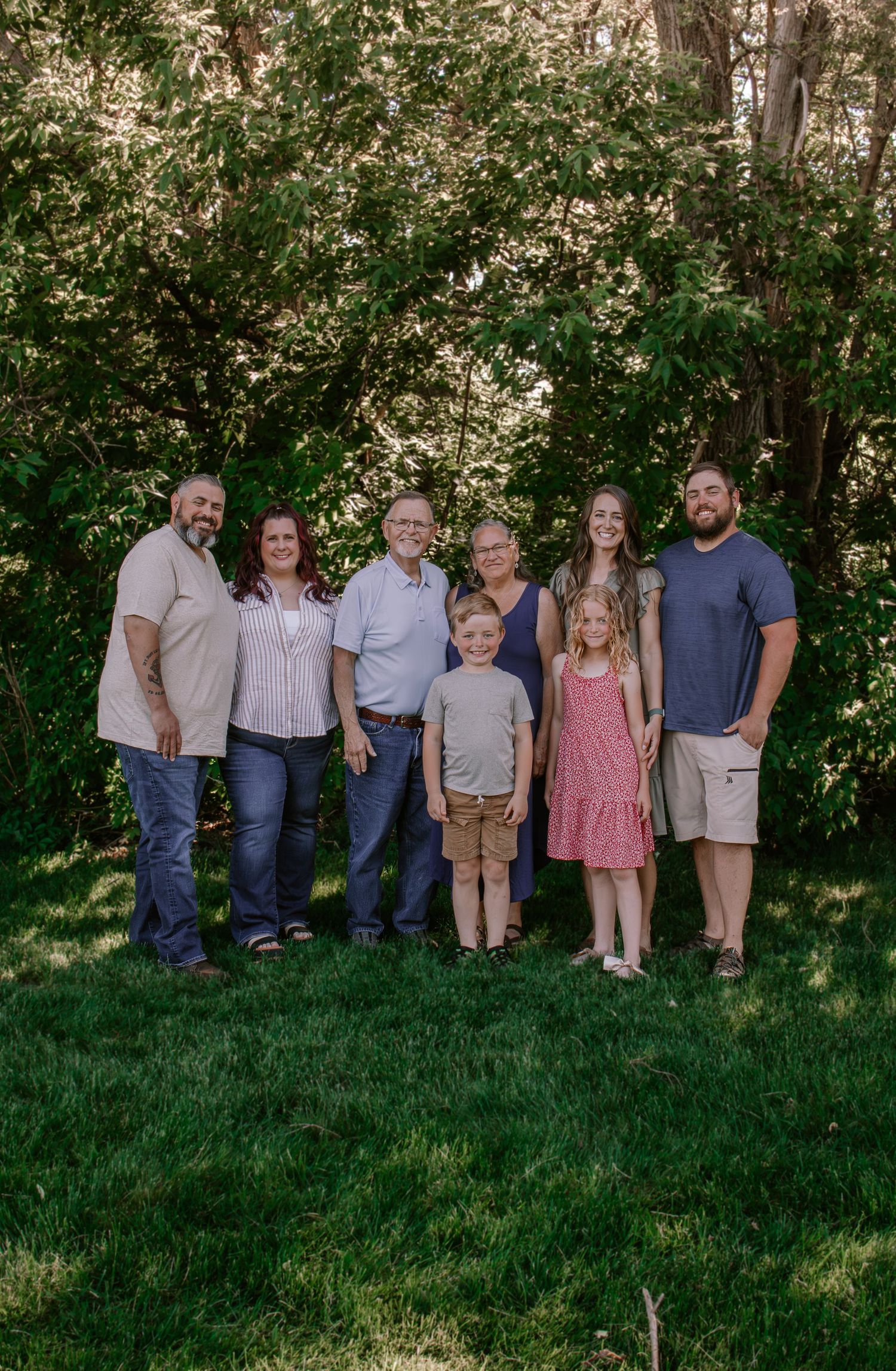 Multi-generational family of eight smiling together outdoors on green grass with lush trees in background.