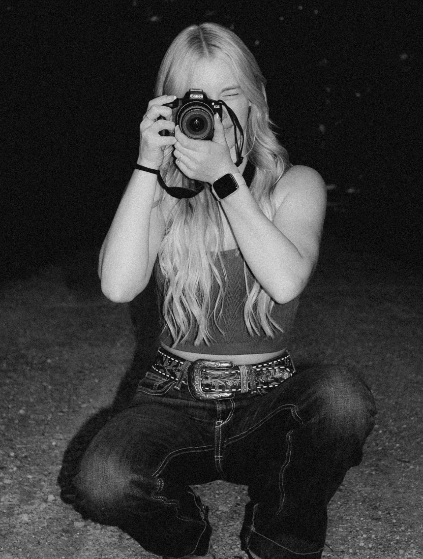 Black and white photo of a photographer crouched down taking a picture with a DSLR camera, wearing jeans and a studded belt.