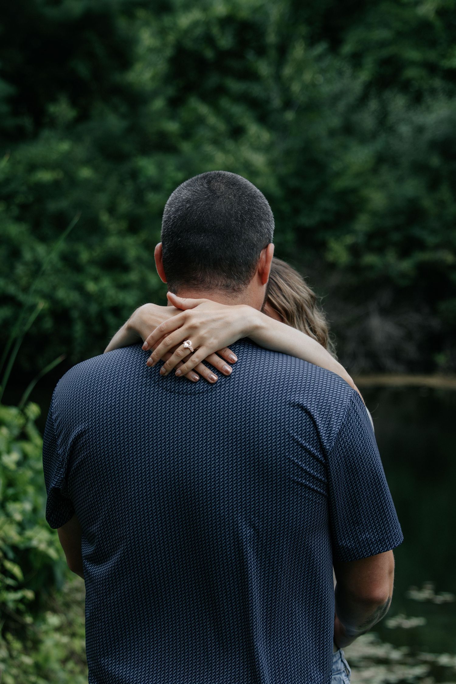 Couple embracing outdoors near a pond, woman with engagement ring resting hand on man's shoulder.