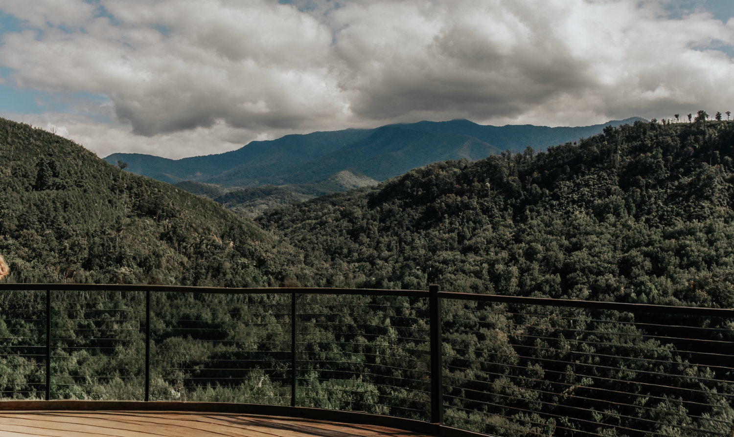 A panoramic view of green mountainous landscape with dramatic cloudy skies above dense forest terrain.