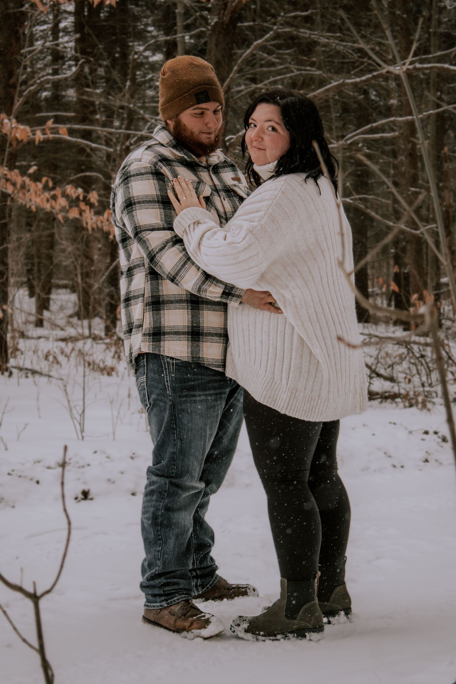 A couple embraces in a snowy winter forest scene wearing cozy sweaters and casual winter attire.
