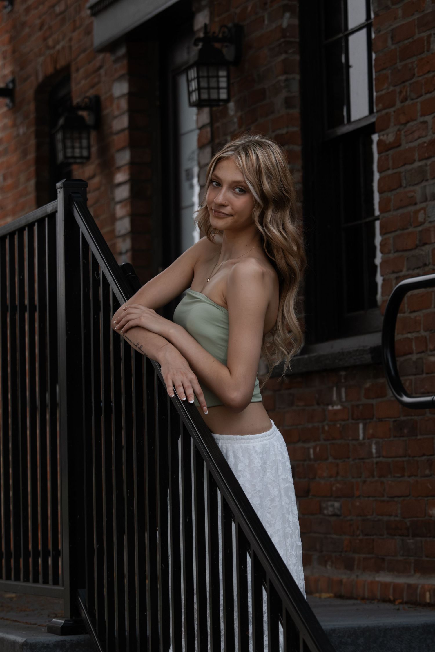 Young woman in sage green tube top and white skirt posing on urban brick building staircase.
