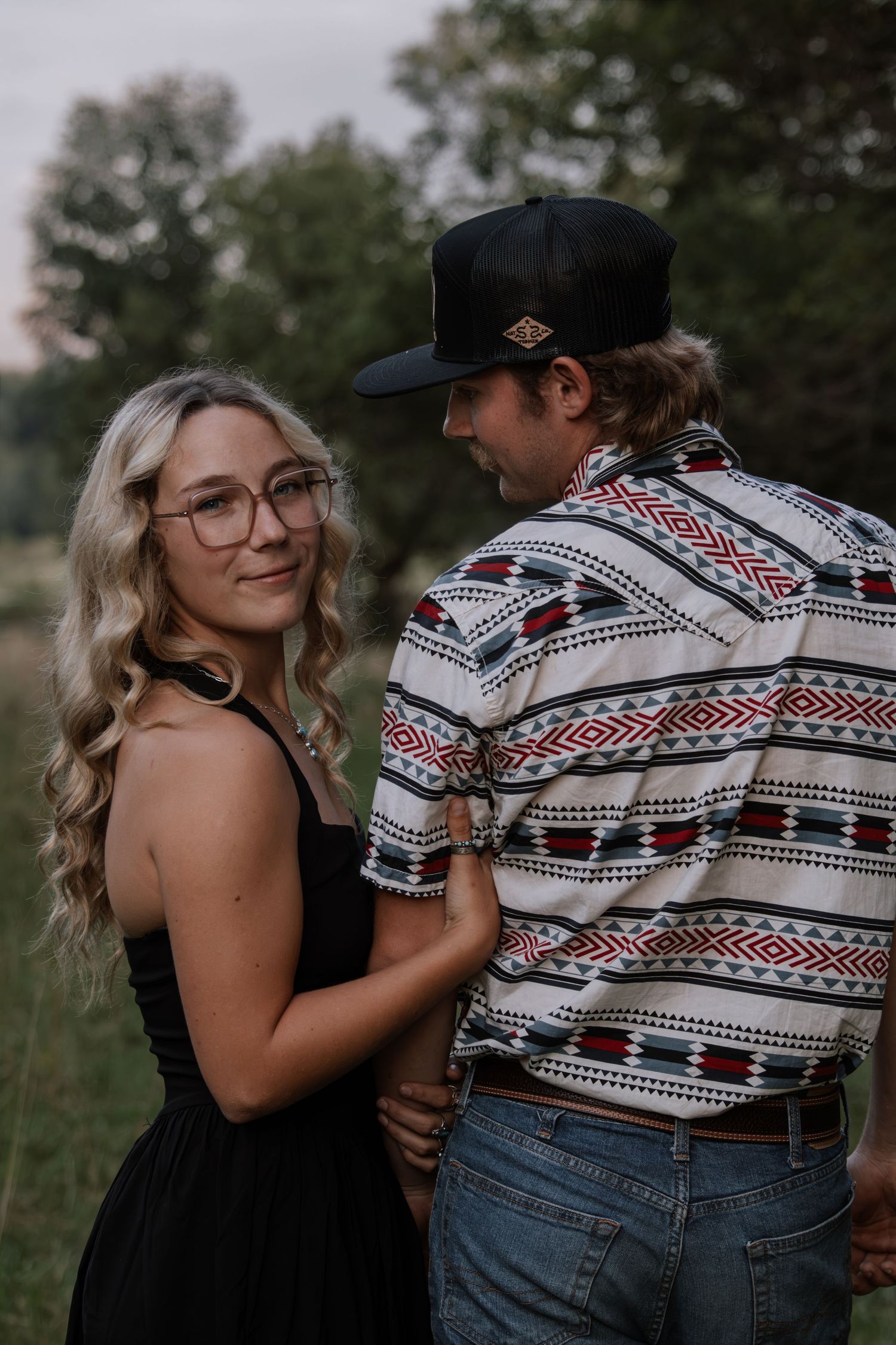 A blonde woman in a black dress and glasses smiles while a man in a patterned shirt and black cap holds her hands outdoors.