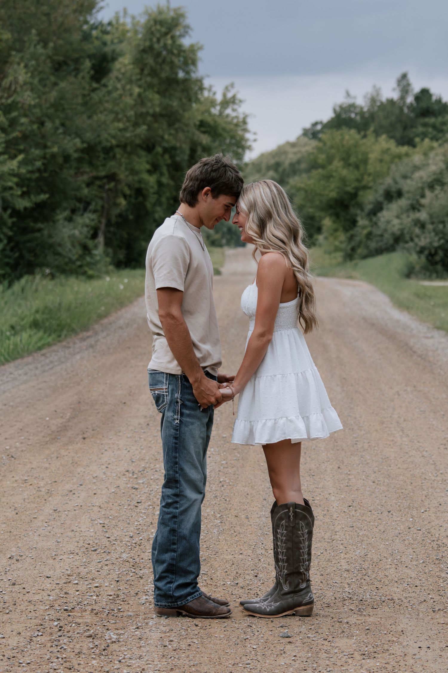 Couple holding hands on a dirt road, woman in white dress and cowboy boots, man in jeans and beige shirt.