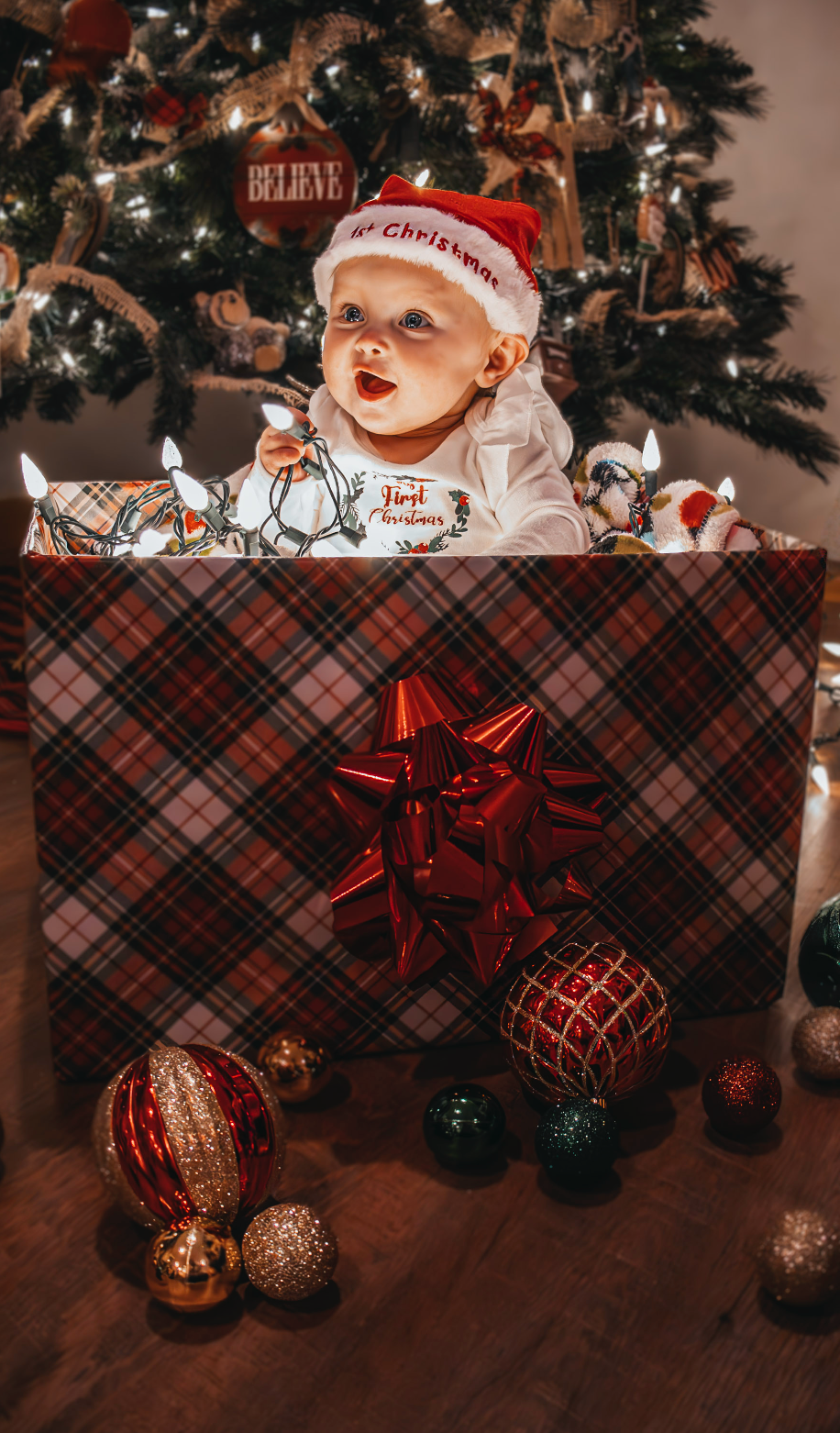 Red and gold Christmas ornaments arranged on plaid fabric beneath a decorated Christmas tree in warm lighting.
