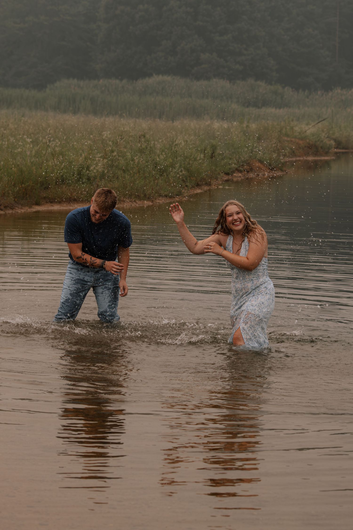 Couple laughing and splashing in a shallow river, wearing jeans and a light blue dress, with green reeds in the background.