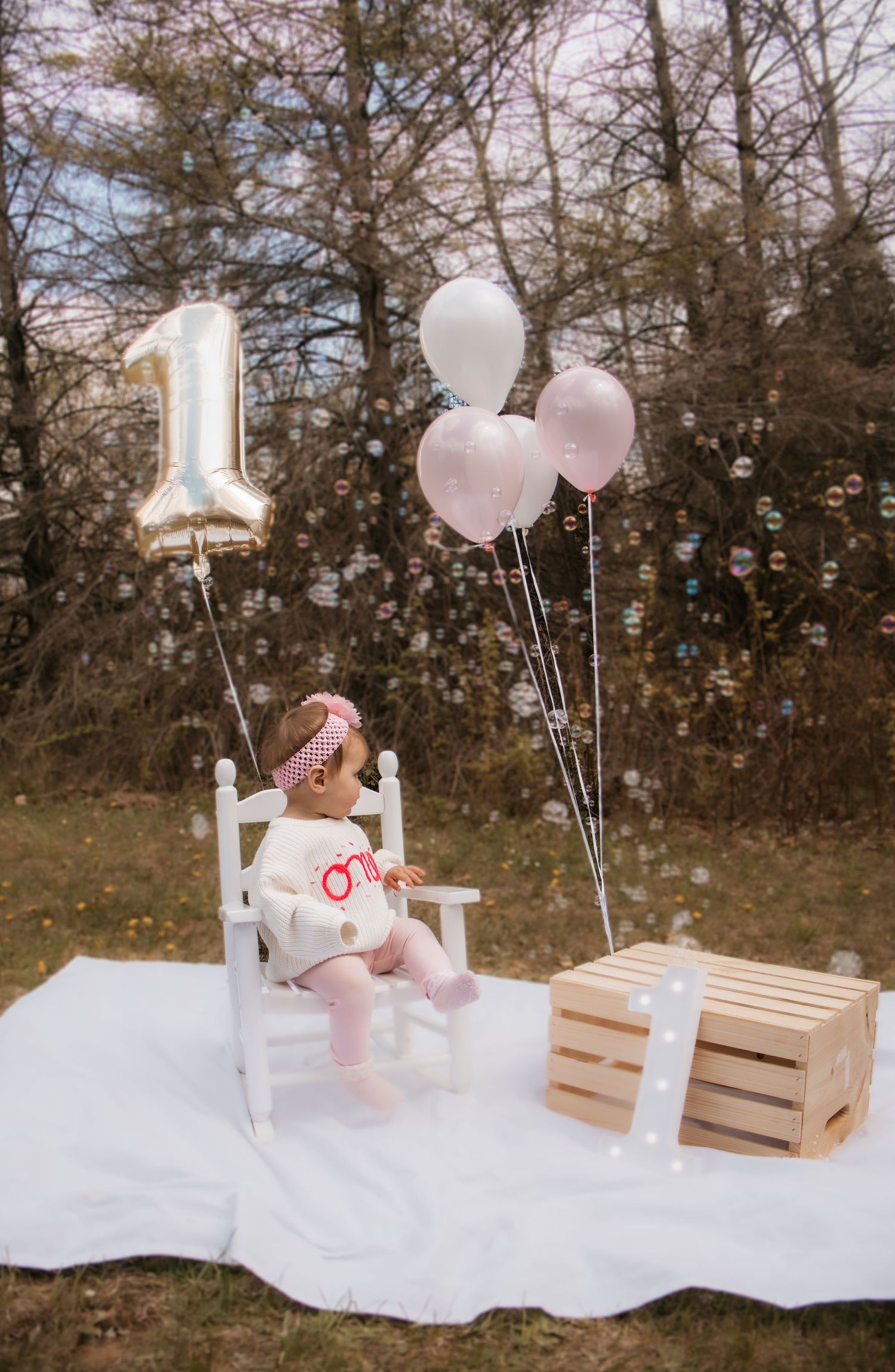 Baby girl's first birthday outdoor photo with number 1 balloon, pink balloons, and bubbles floating in the air.
