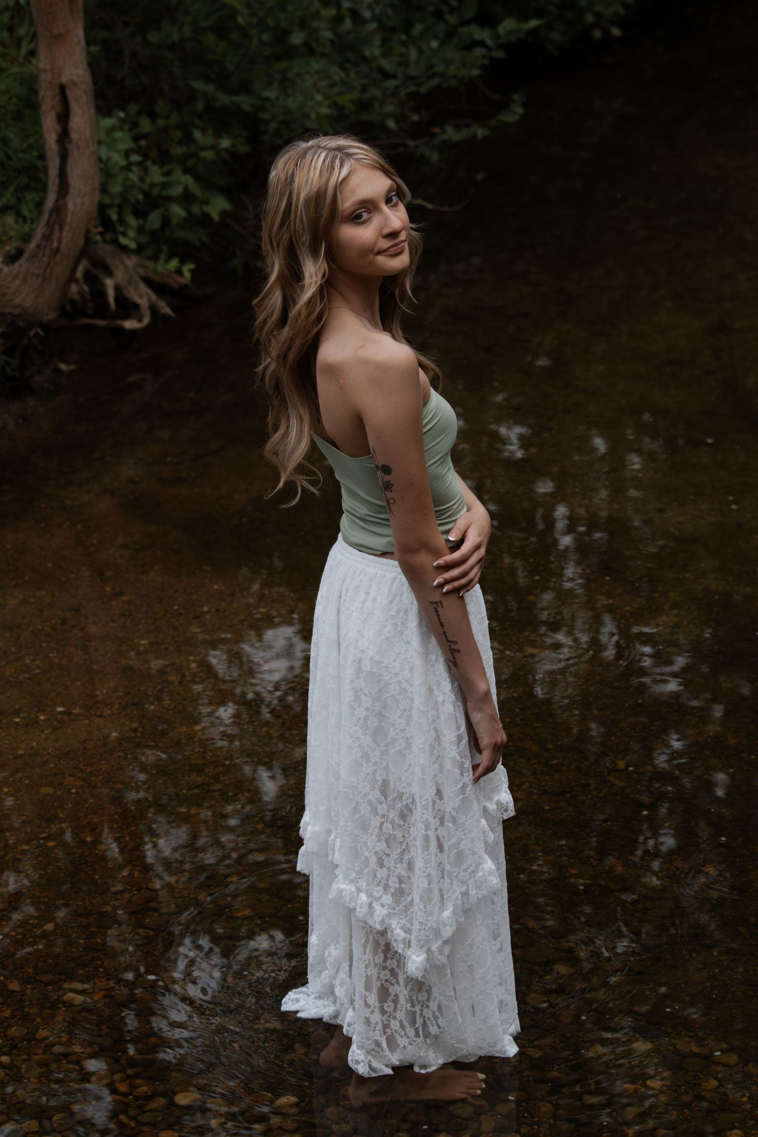Blonde woman in green top and white lace skirt standing barefoot in a shallow creek.