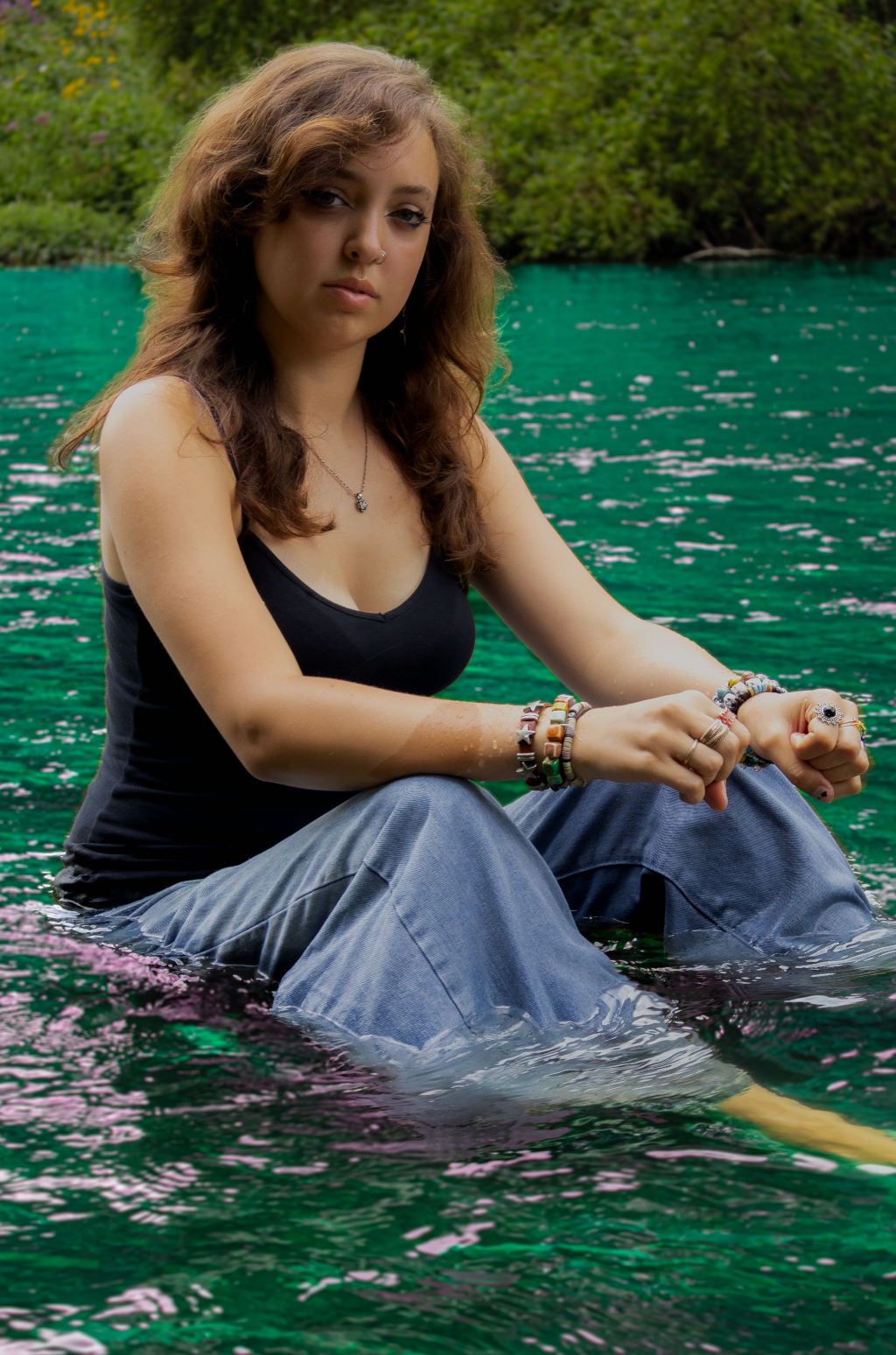 Figure in a black tank top and blue jeans sits at the edge of vibrant turquoise water on a summer day.