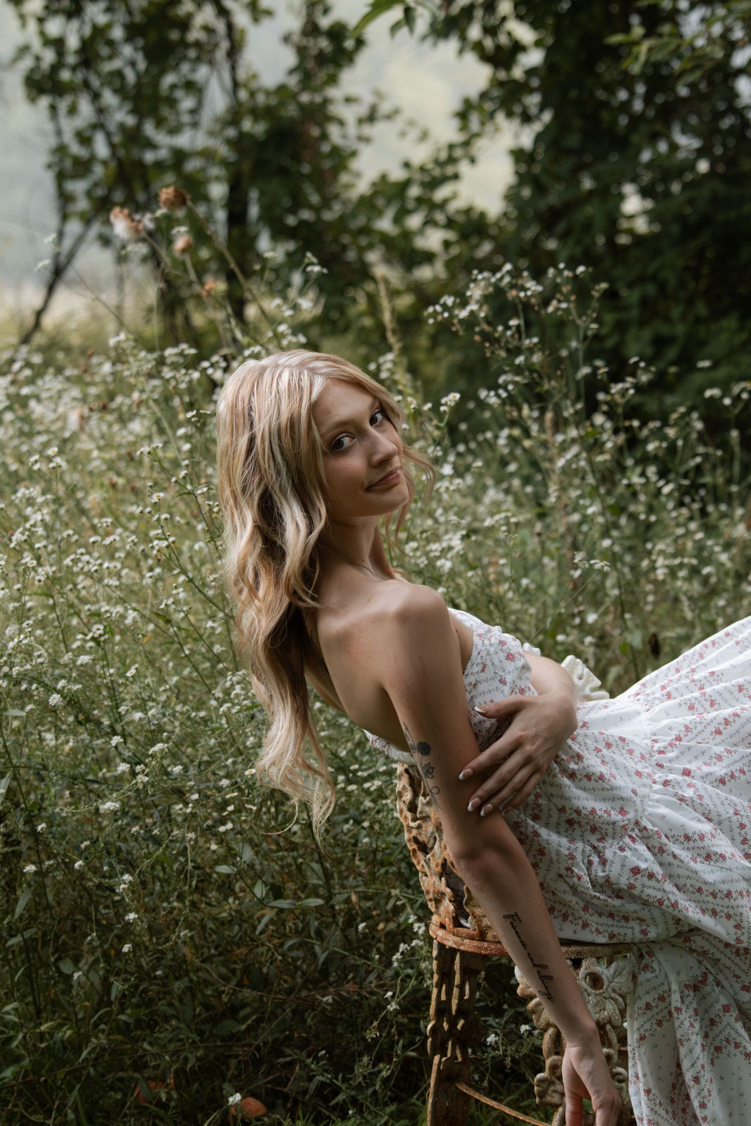 Blonde woman in floral dress sitting among wildflowers in a lush green garden, smiling softly at camera.