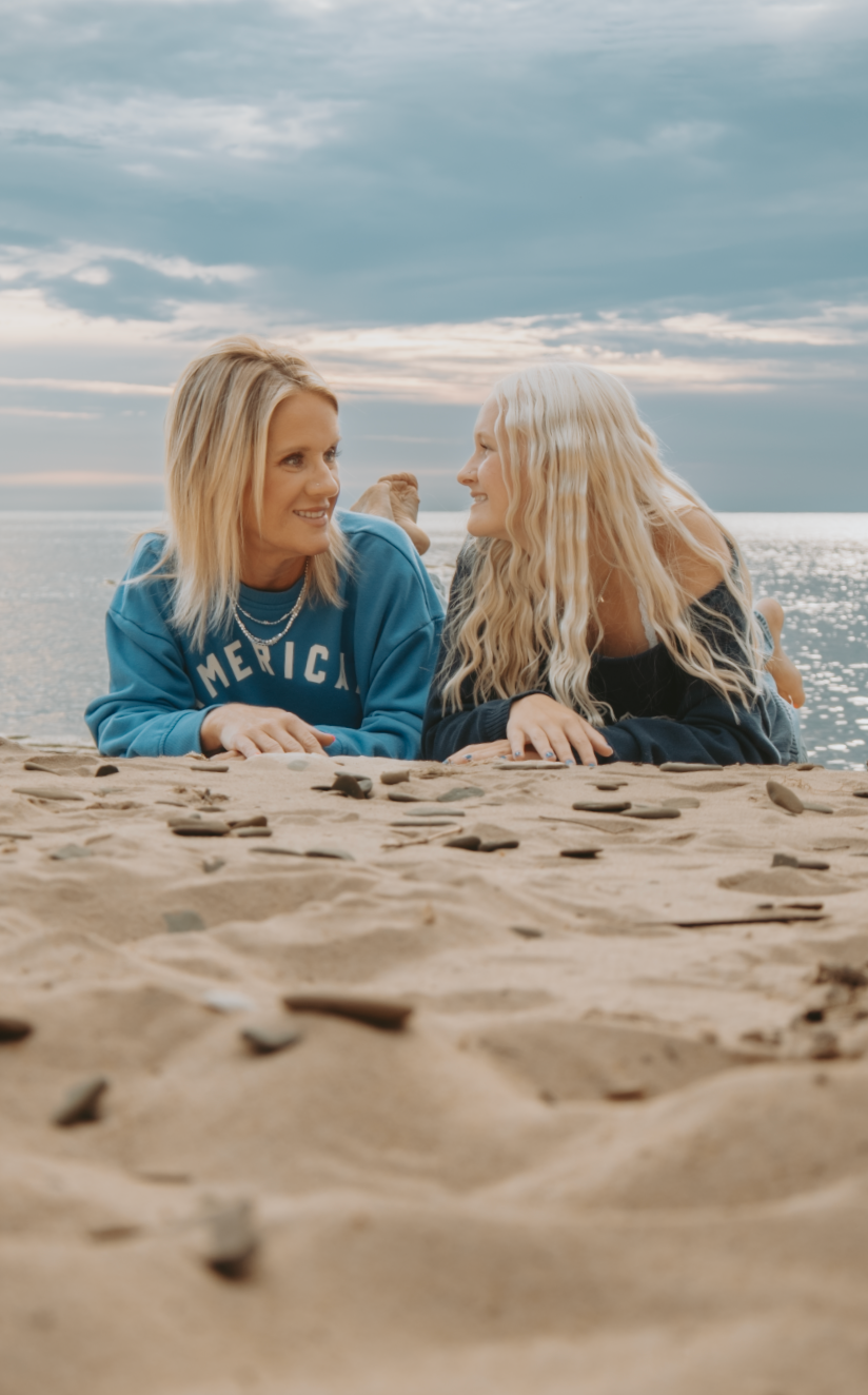 Two friends lying on sandy beach wearing blue sweatshirts share laughter under cloudy sky at the shoreline.