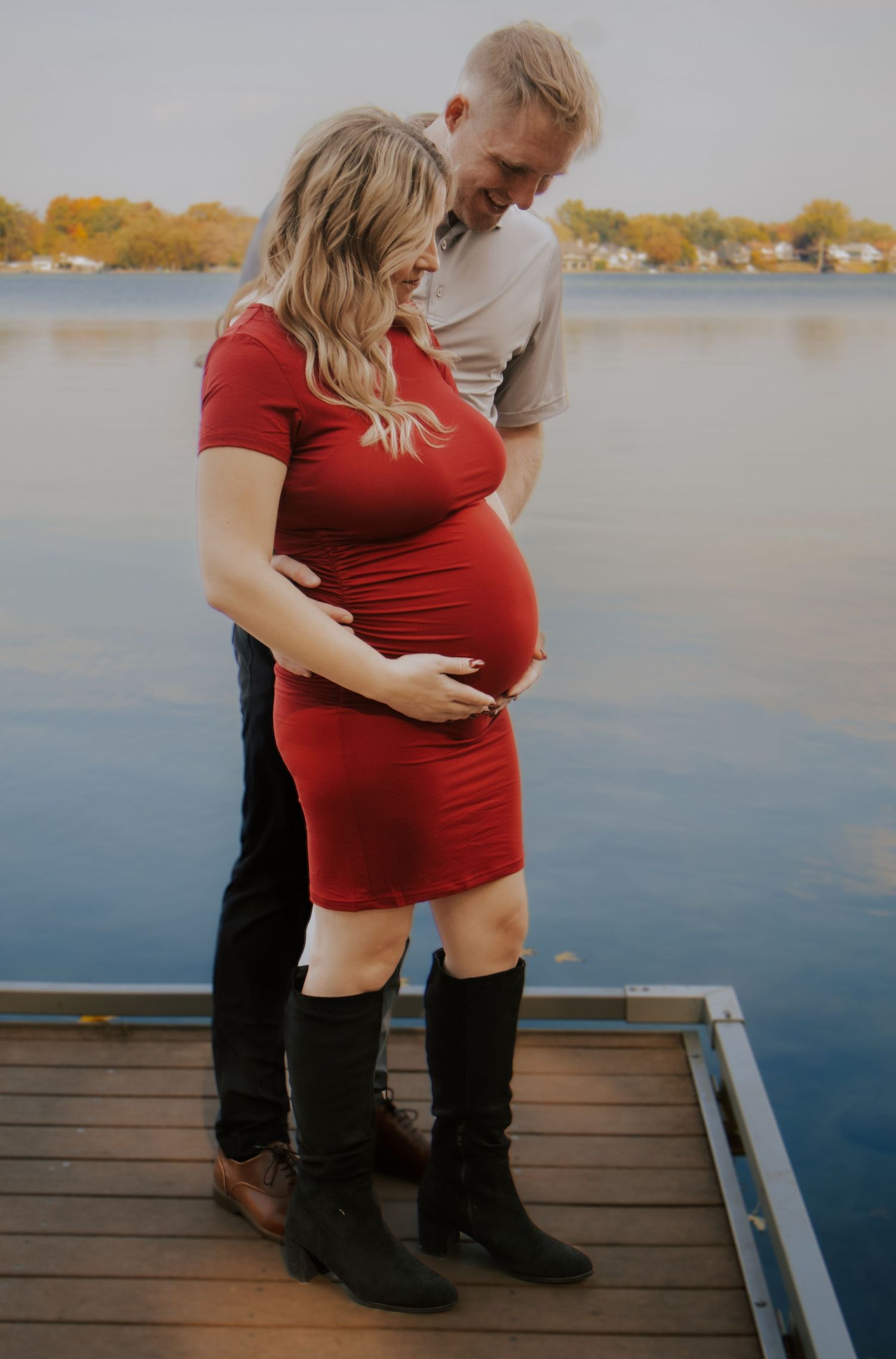 Maternity photos on wooden dock with couple in red dress and gray shirt by calm lake.