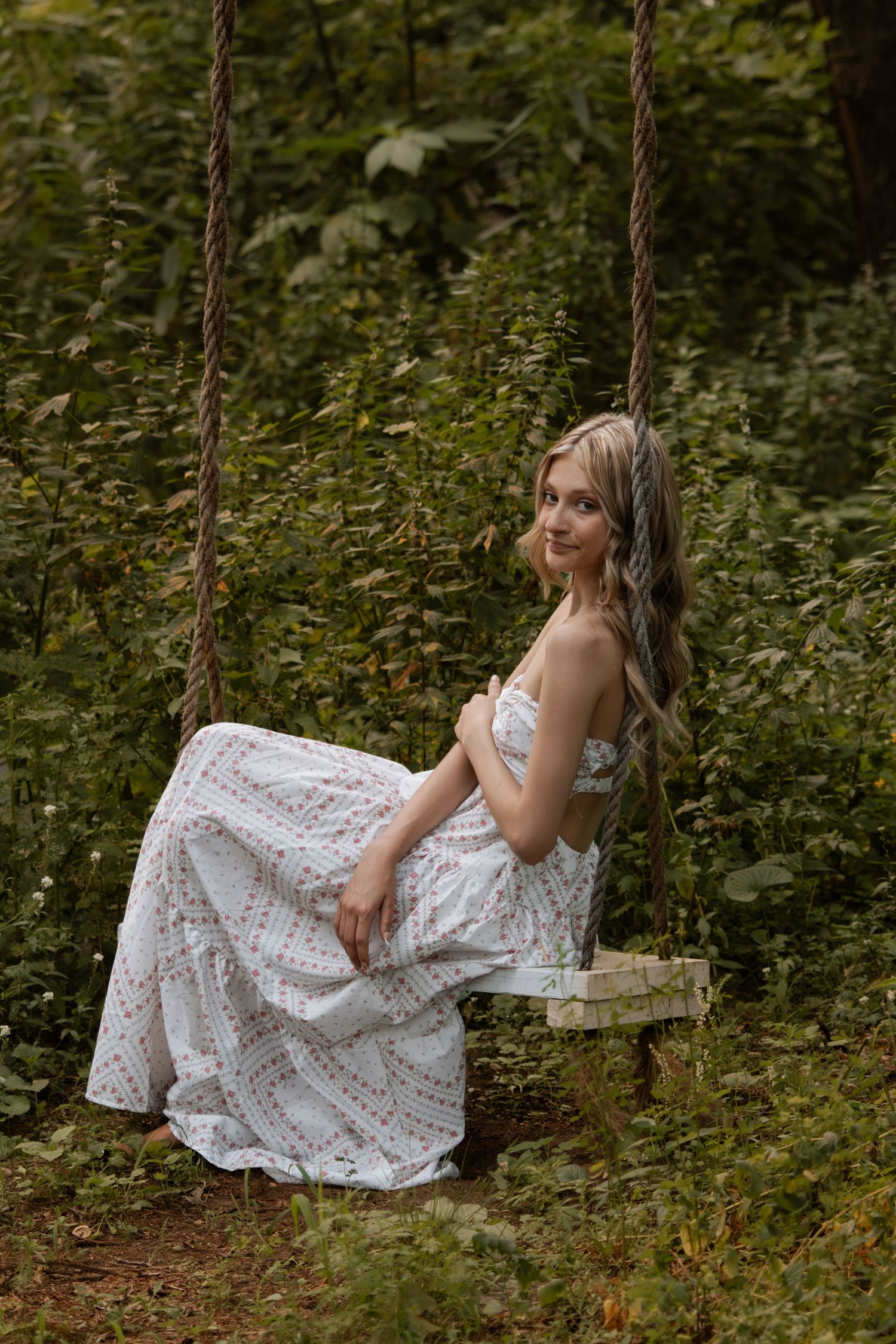 Young blonde woman in white floral maxi dress sitting gracefully on a rope swing surrounded by lush green forest foliage.
