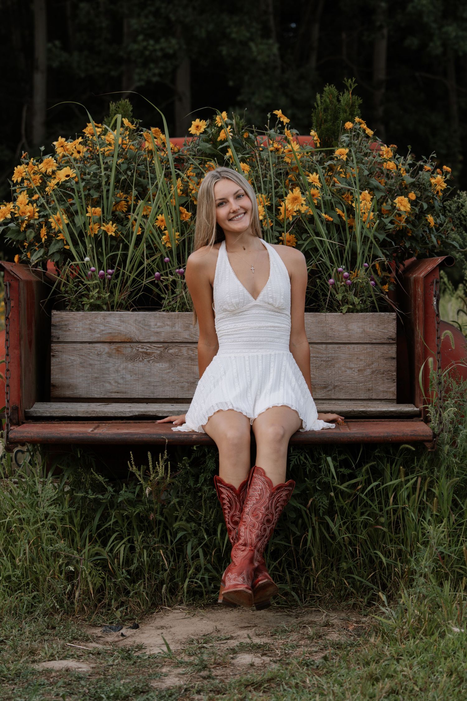 Smiling blonde woman in white halter dress and red cowboy boots sitting on rustic bench with yellow flowers.