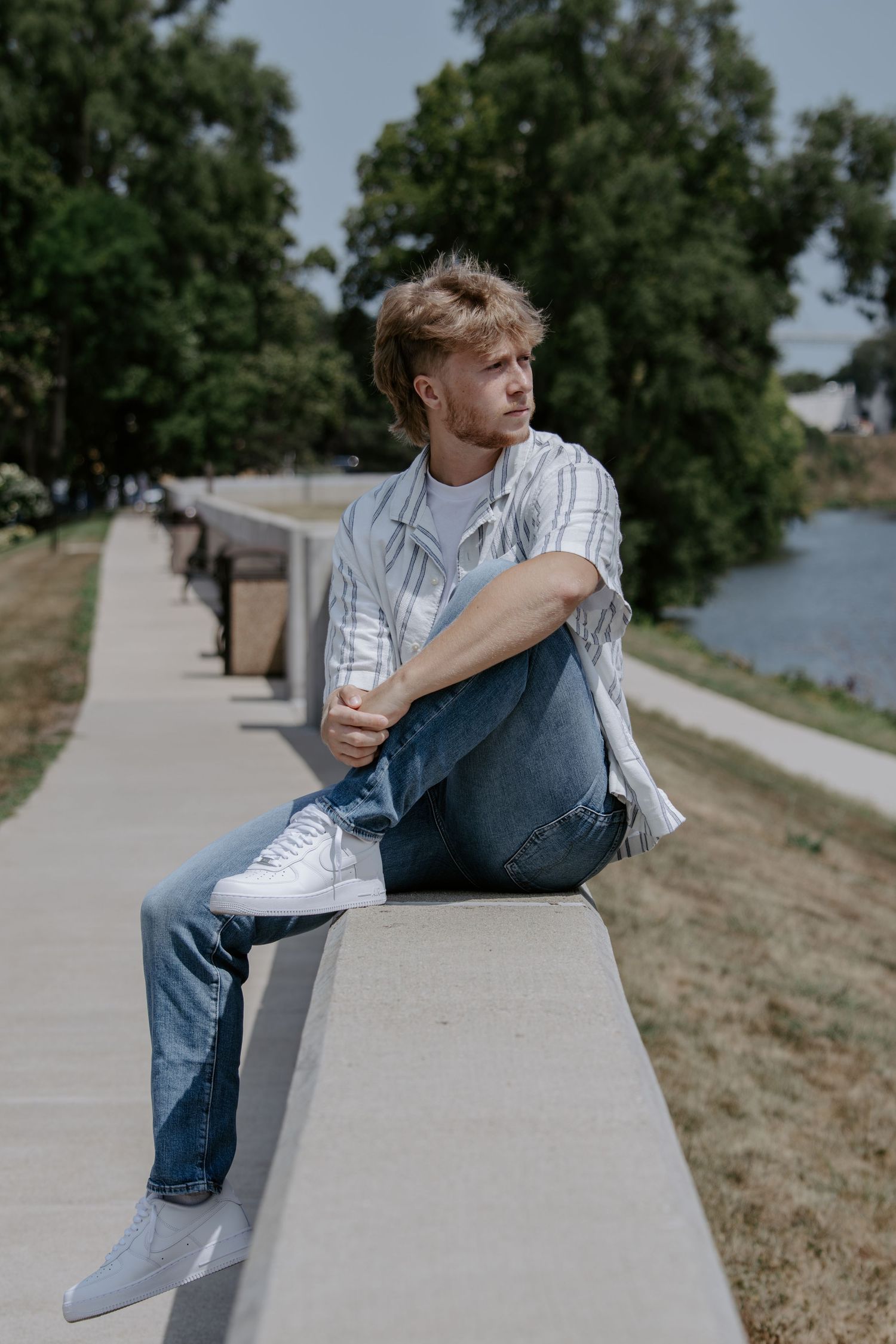 Young man in jeans and striped shirt sitting on a concrete ledge in a sunny park near a river.