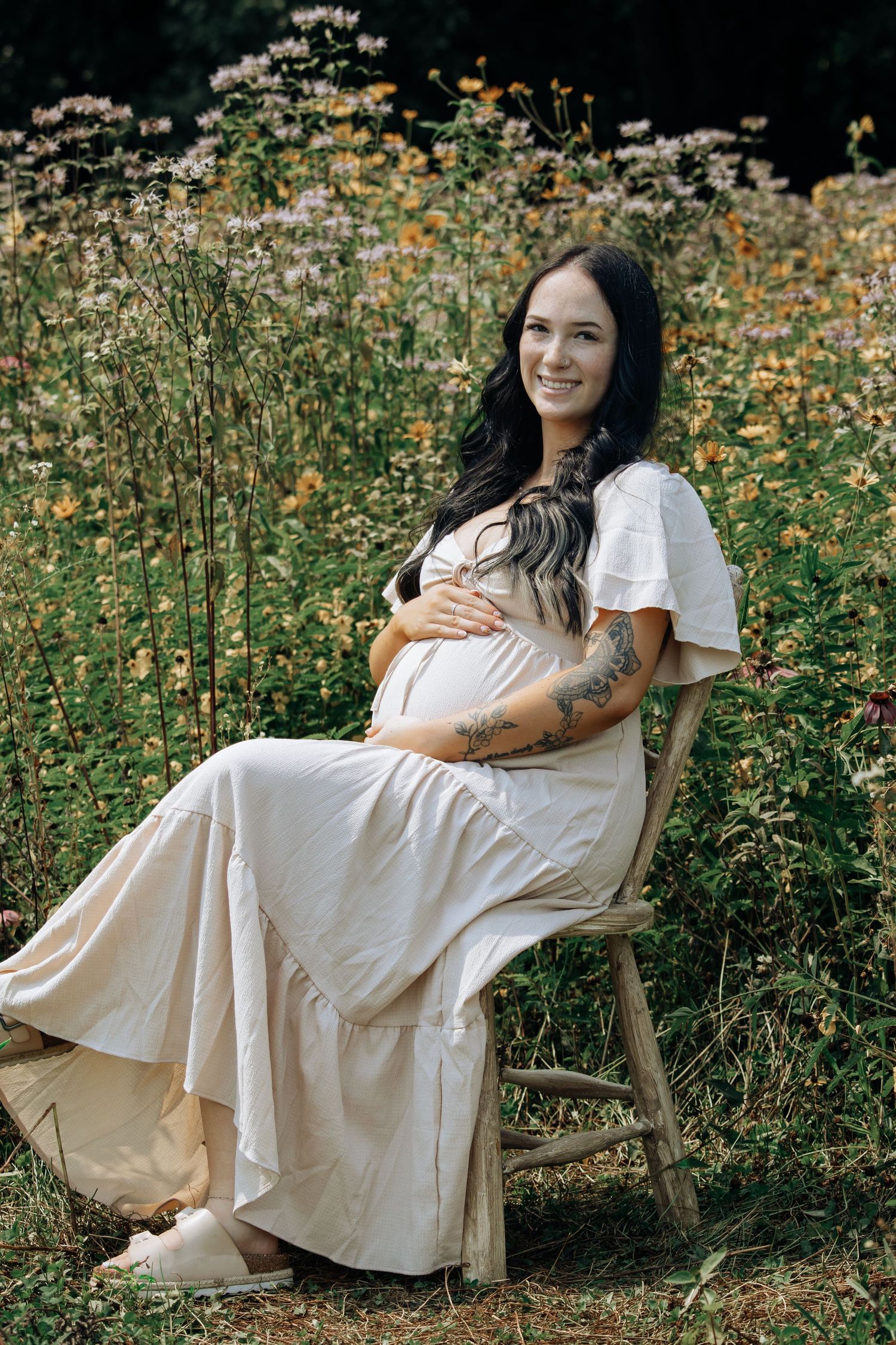 Smiling pregnant woman with tattoos in blush maxi dress sits on wooden chair surrounded by wildflowers.