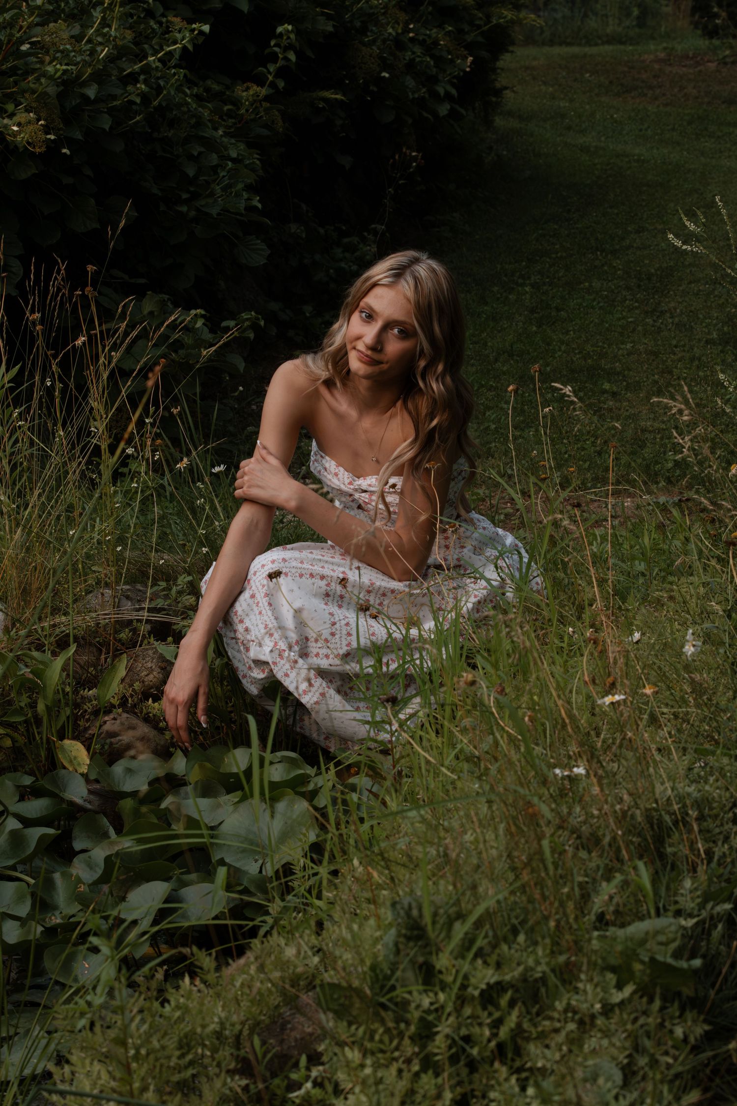 A young blonde woman in a floral sundress sits gracefully among wildflowers and lush green grass in a serene forest setting.