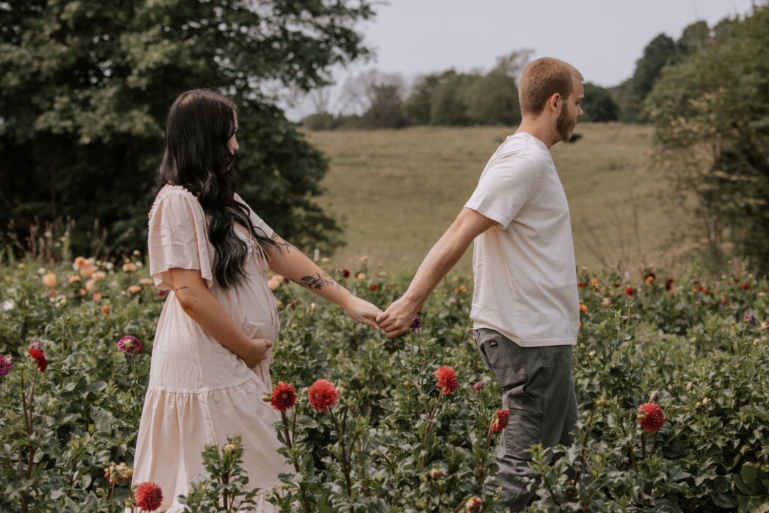 Pregnant woman in white dress holds hands with man in flower field.