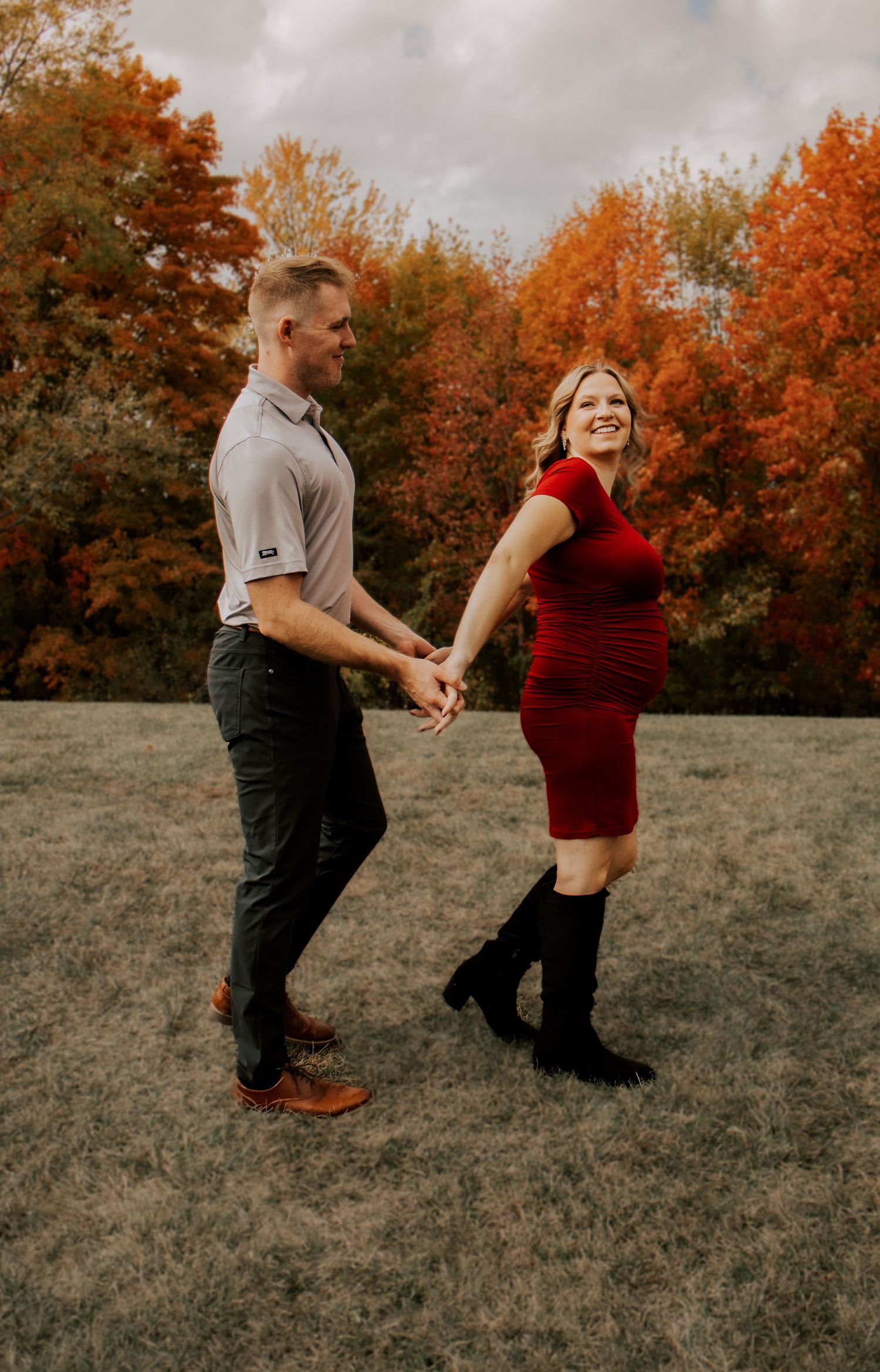 A playful couple dances together in a field with autumn trees in the background.