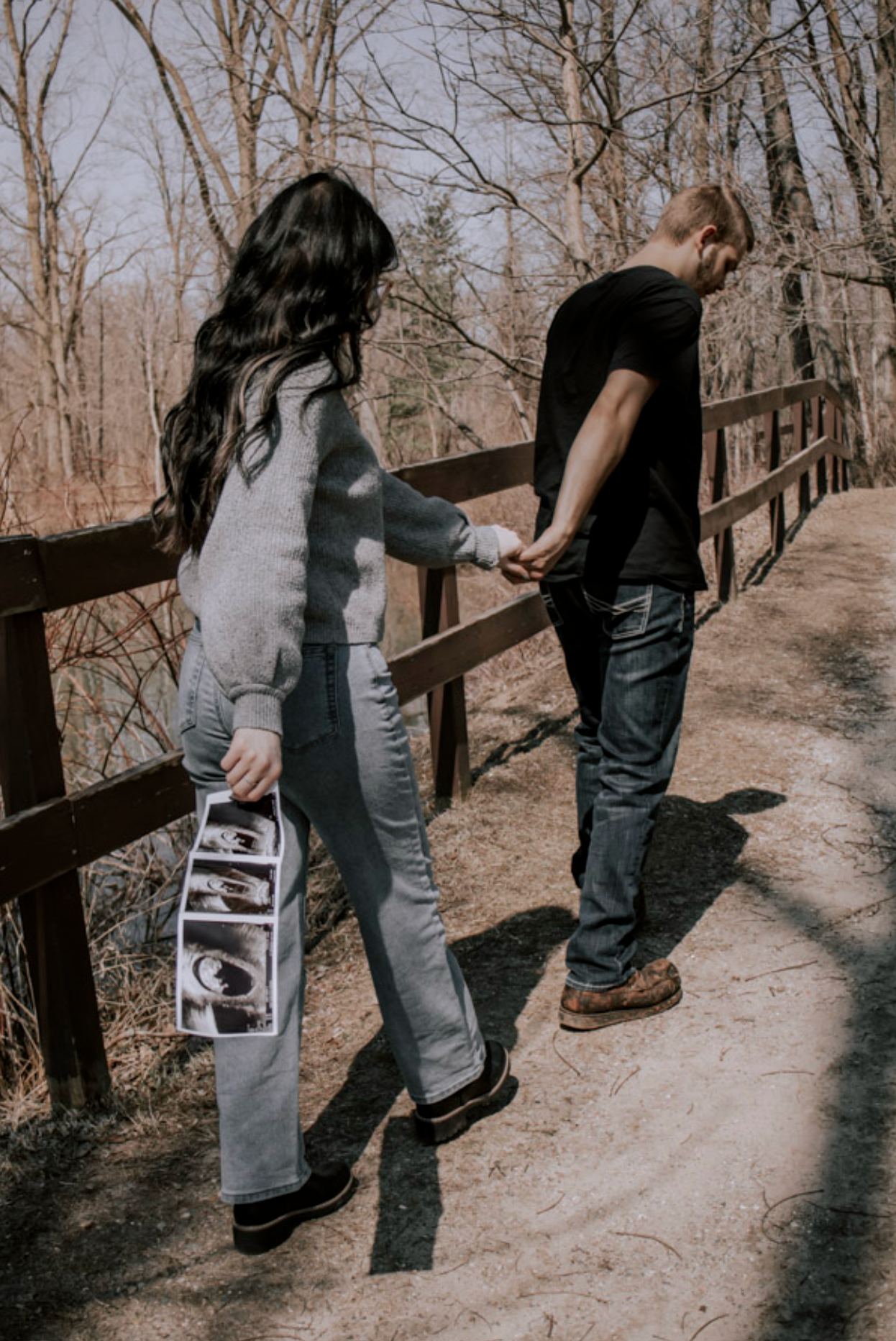 Couple holding hands on a wooden bridge in a forest, woman holding ultrasound photos, pregnancy announcement outdoors.