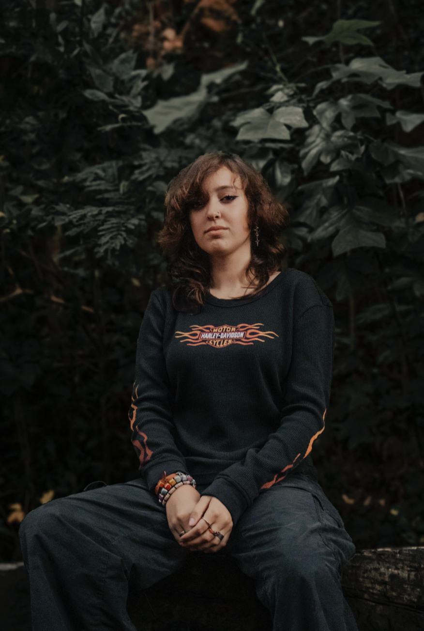 Someone wearing a black sweater with orange accents sits casually on a wooden bench in a moody garden setting.