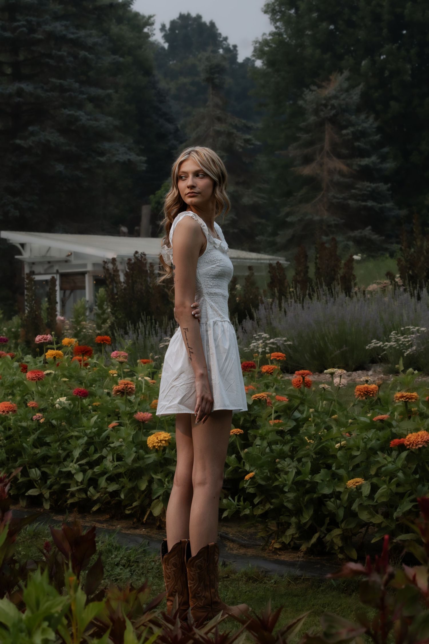 Young woman in white dress and cowboy boots standing in a colorful flower garden with evergreen trees in background.