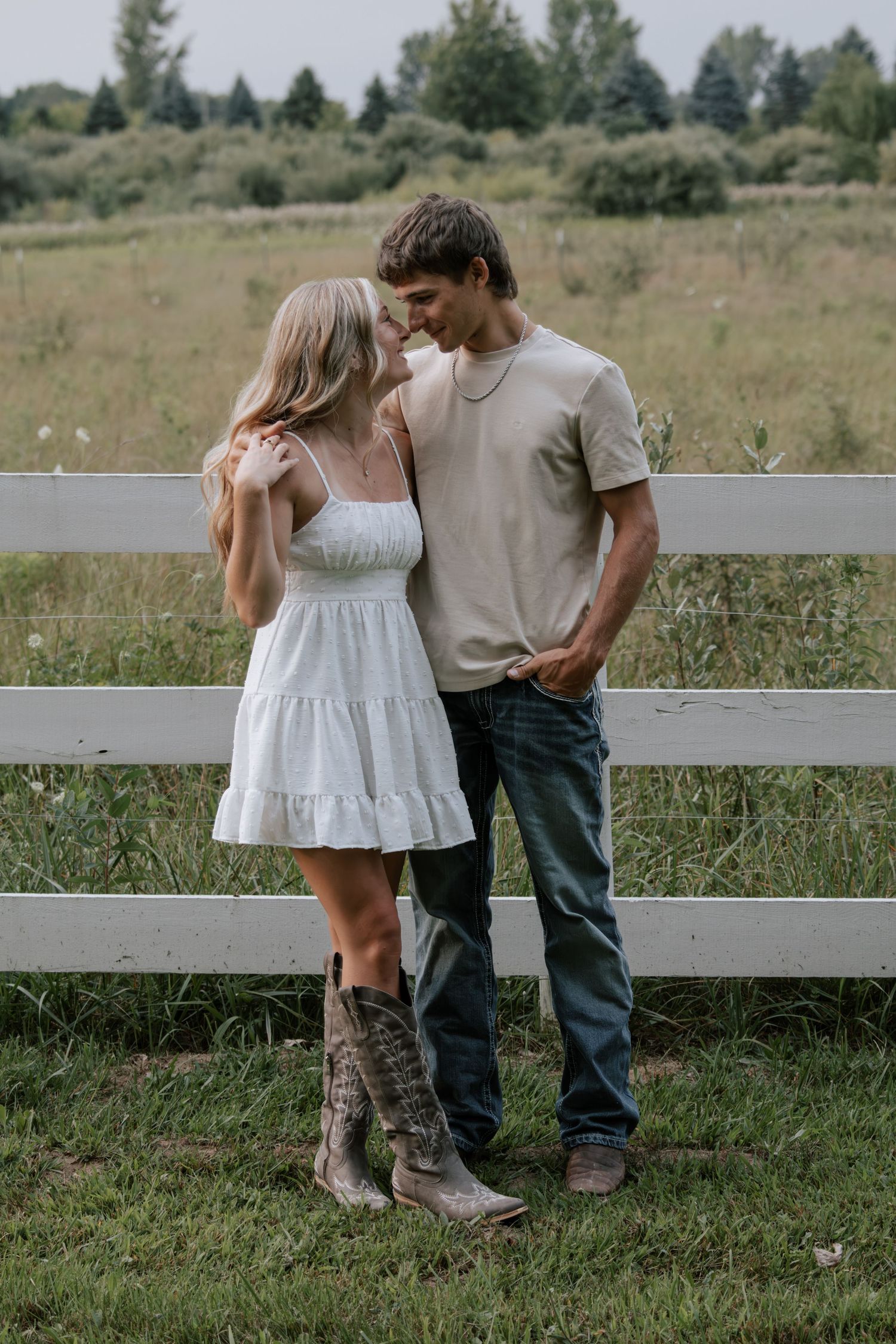 Young couple in romantic pose by white fence, woman in white dress and cowboy boots, man in beige shirt and jeans.