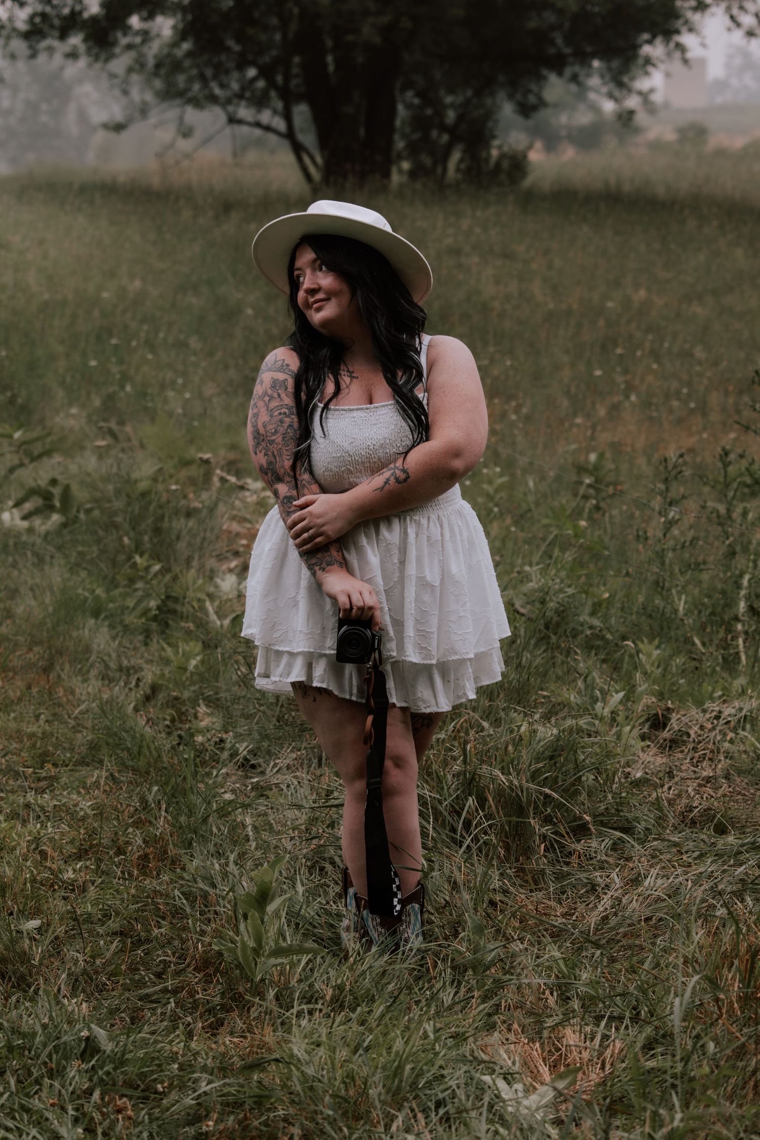 Woman in white dress and hat holds camera in a lush green field, showcasing tattoos and a warm smile.