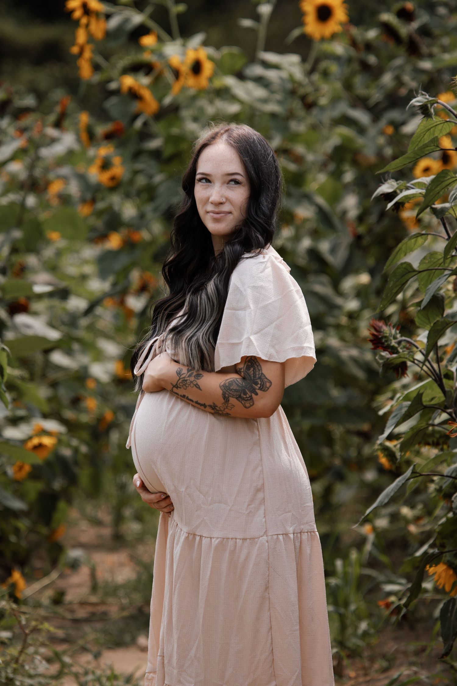A pregnant woman in a blush dress cradles her baby bump surrounded by sunflowers in a garden.