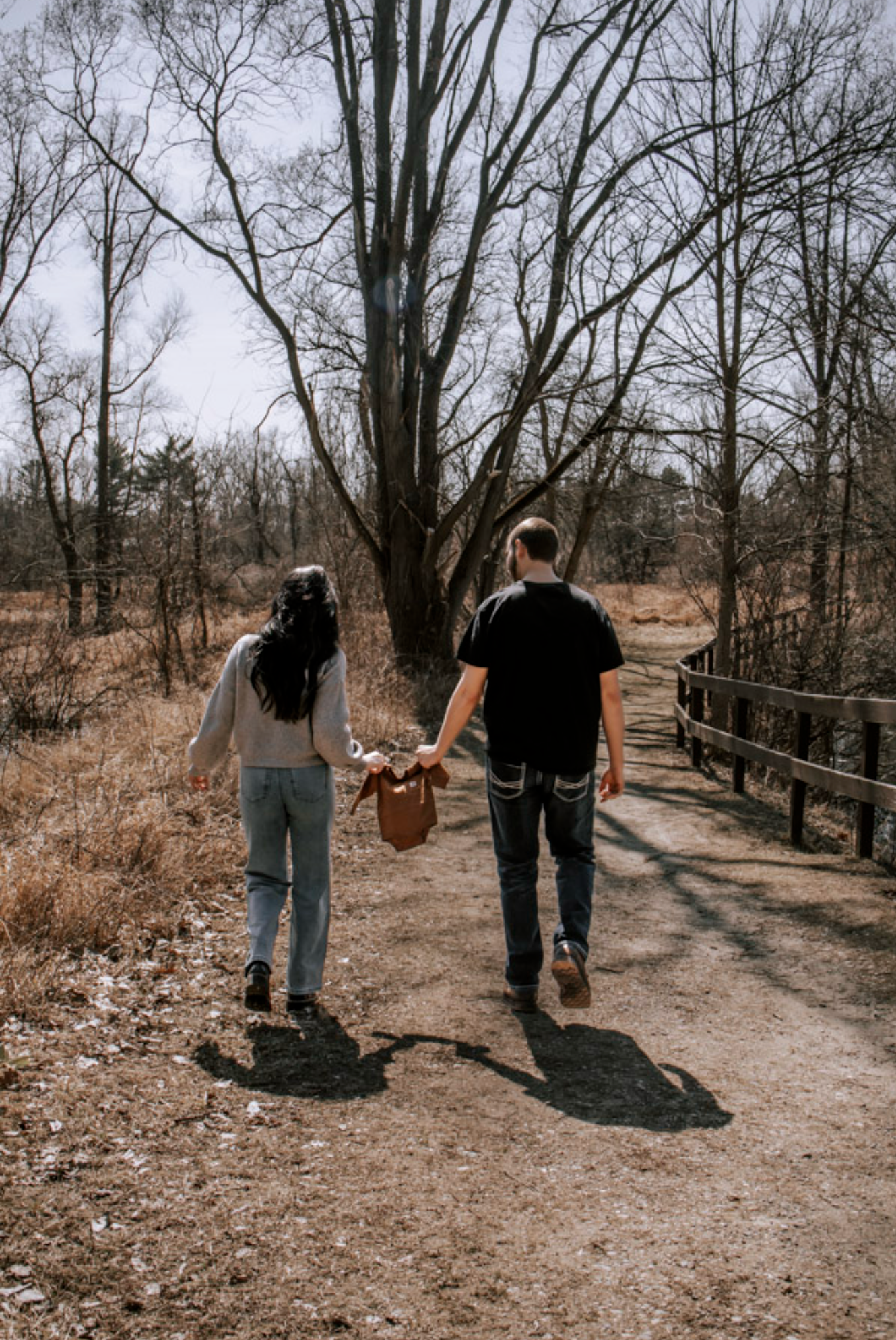 Couple holding hands walking on a dirt path toward a wooden bridge surrounded by bare trees in early spring.