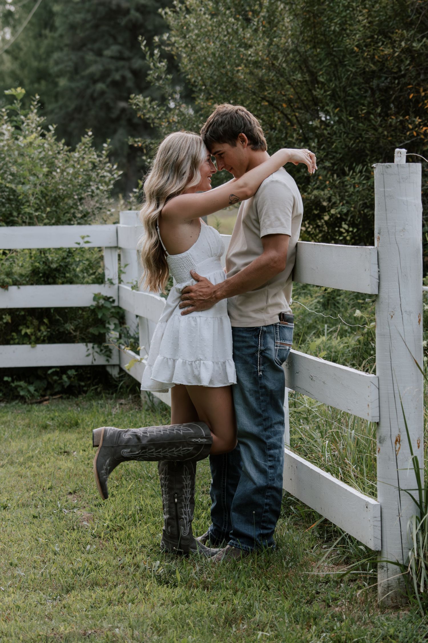 A couple embraces romantically by a white wooden fence in a lush green countryside setting.