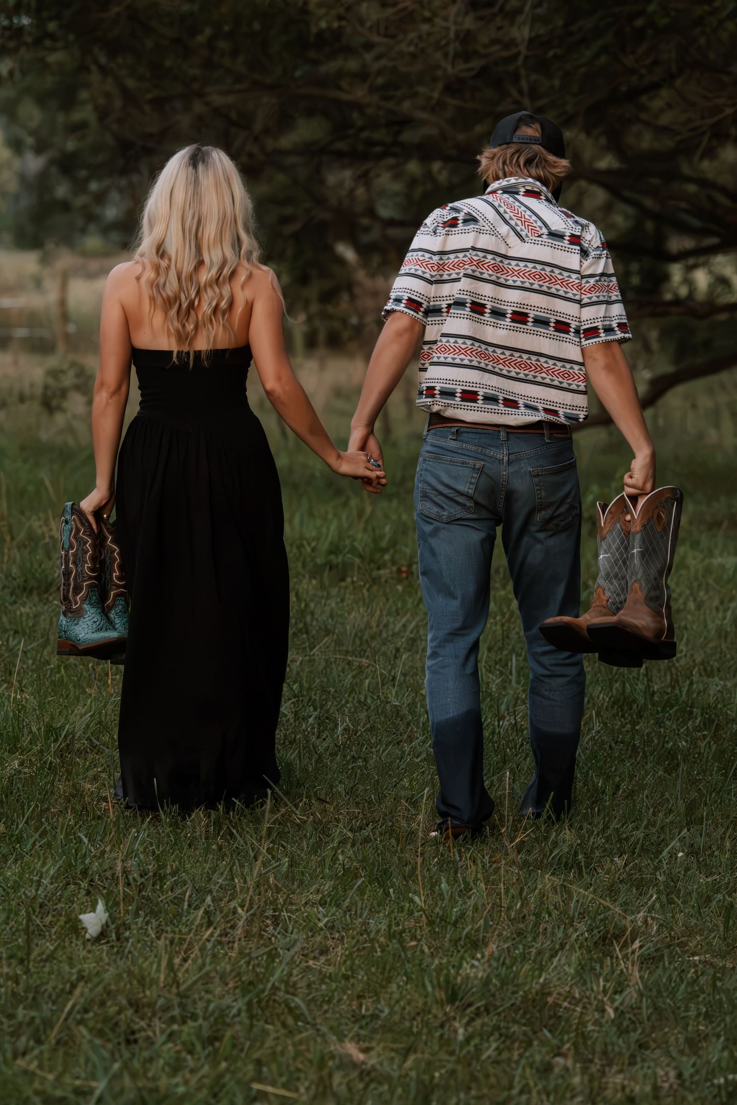 Couple holding hands and cowboy boots walking barefoot through green grass field, woman in black maxi dress, man in patterned shirt.