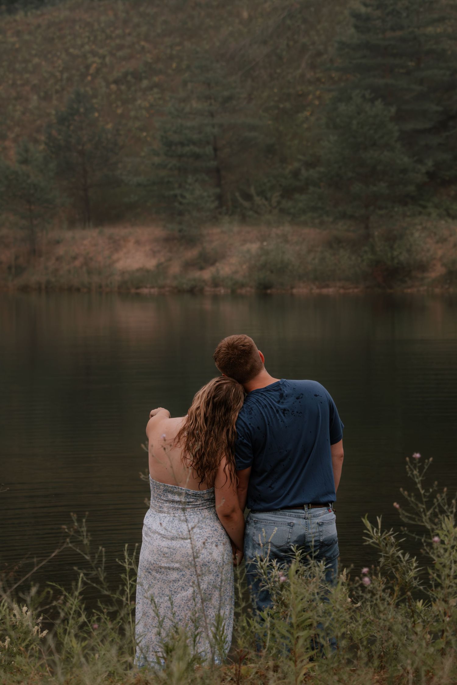 A couple embraces by a serene lake surrounded by autumn trees, viewed from behind in a romantic outdoor setting.