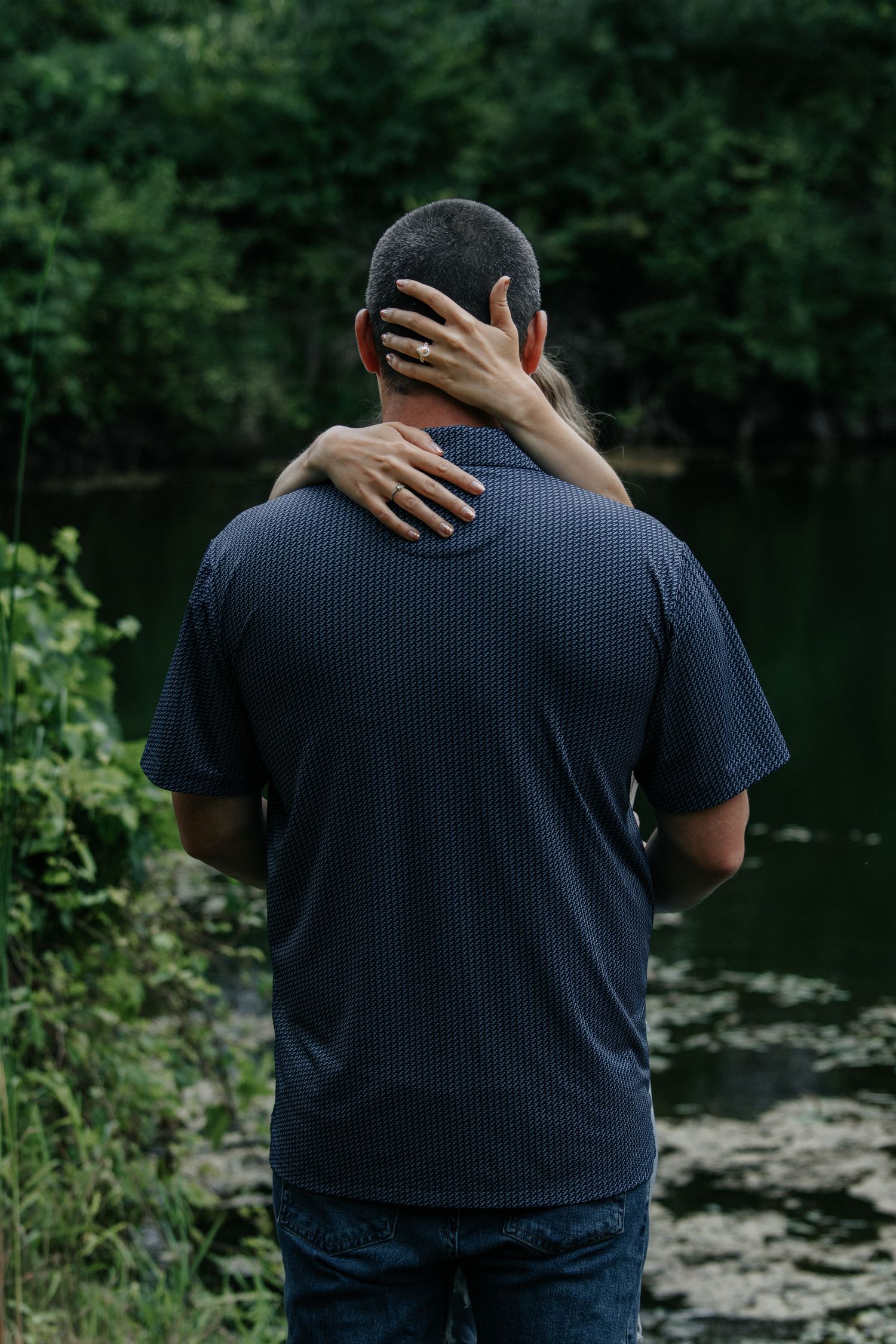 Couple embracing by a serene pond surrounded by lush green trees, viewed from behind.