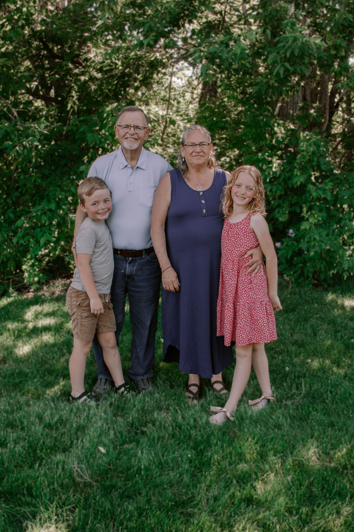 Grandparents smiling outdoors with two young grandchildren on green grass, surrounded by lush trees in a family portrait.