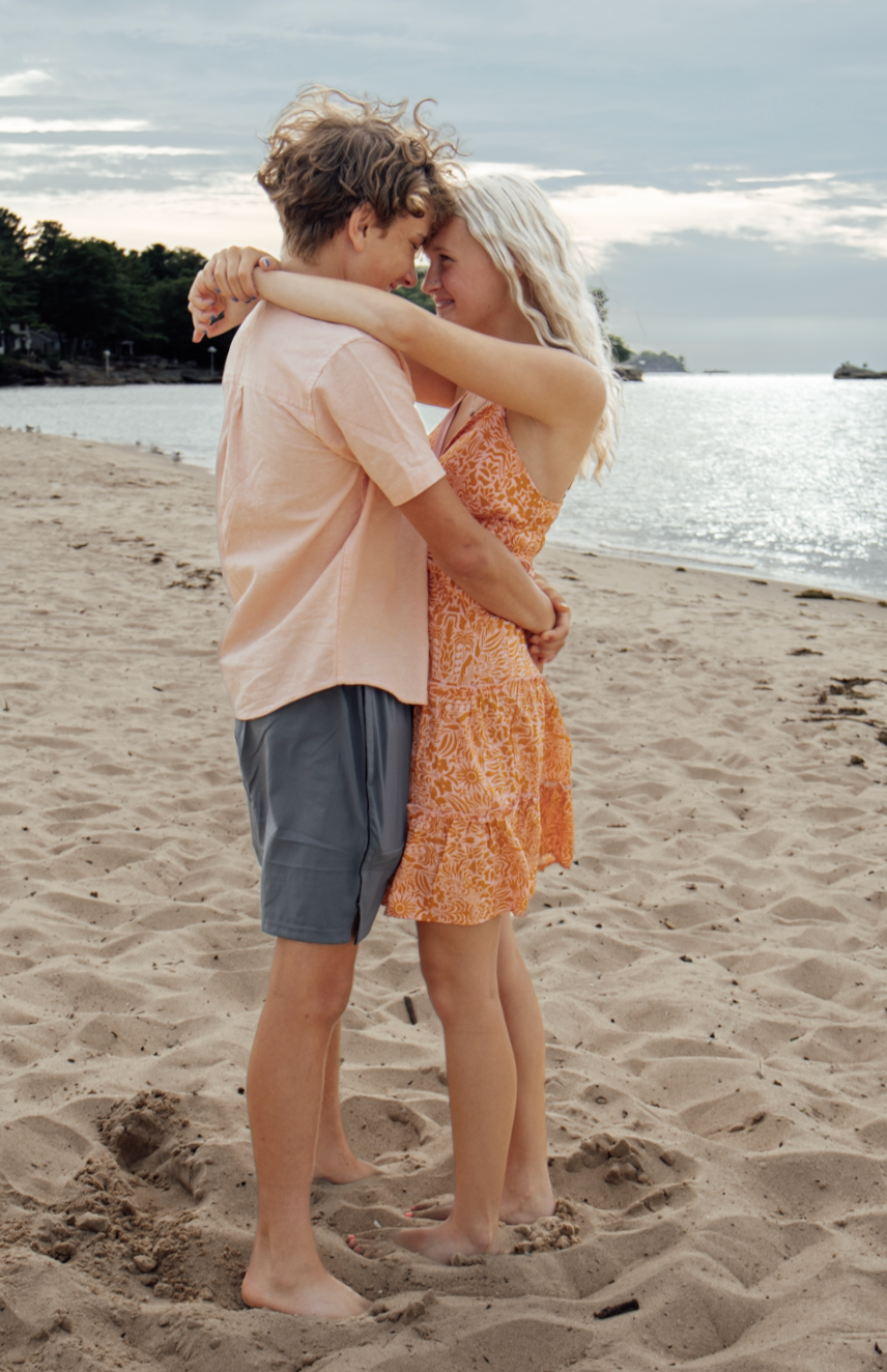 Two small children hug sweetly on a sandy beach with ocean waves and cloudy sky in the background.