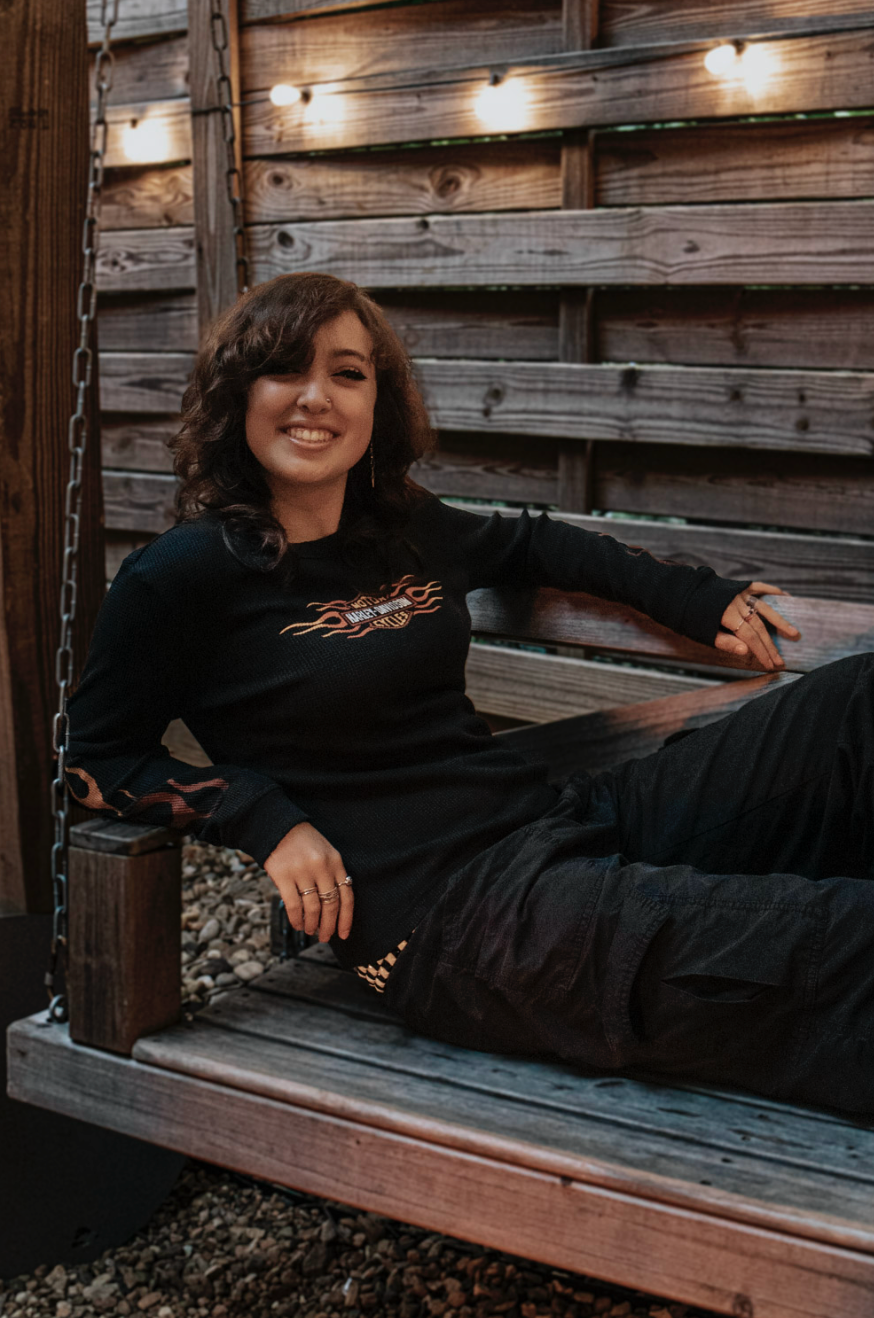 A relaxed figure in a black sweatshirt poses across a rustic wooden bench with string lights decorating the backdrop.